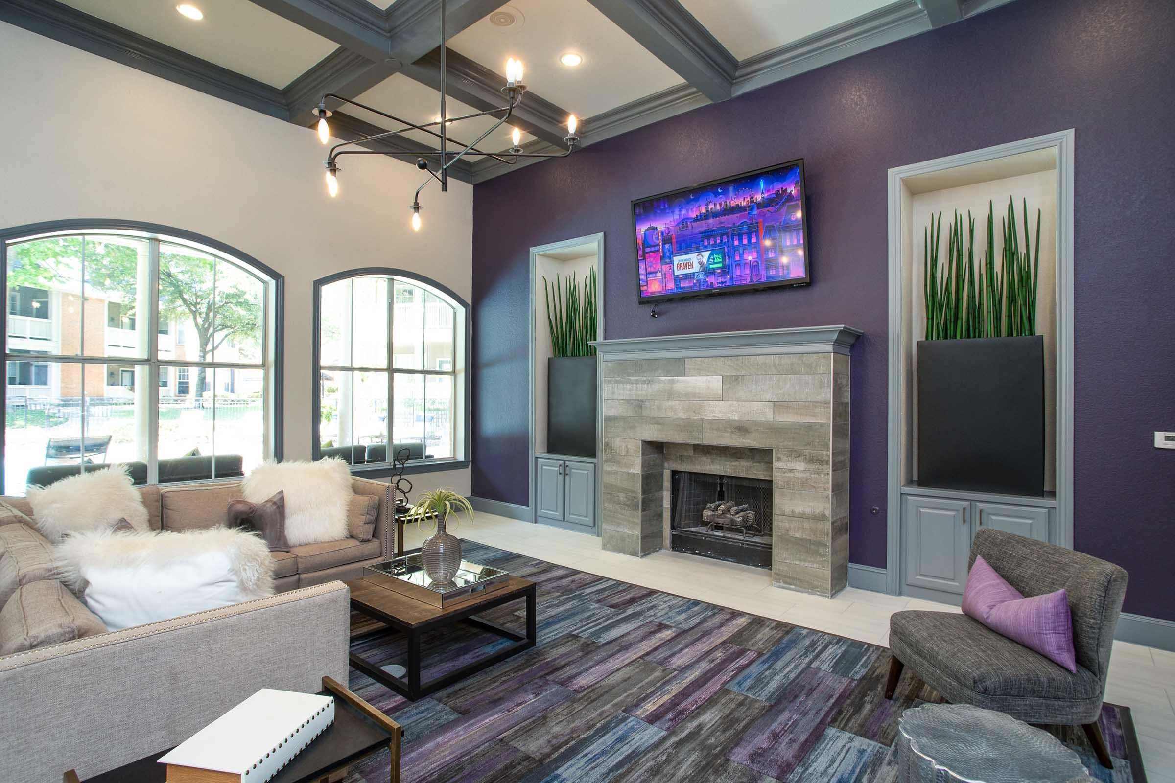 Modern living room featuring a cozy sofa with plush pillows, a coffee table, and a stone fireplace. Large windows allow natural light, and potted plants add greenery. A stylish chandelier and a wall-mounted TV enhance the contemporary decor, complemented by a colorful area rug.