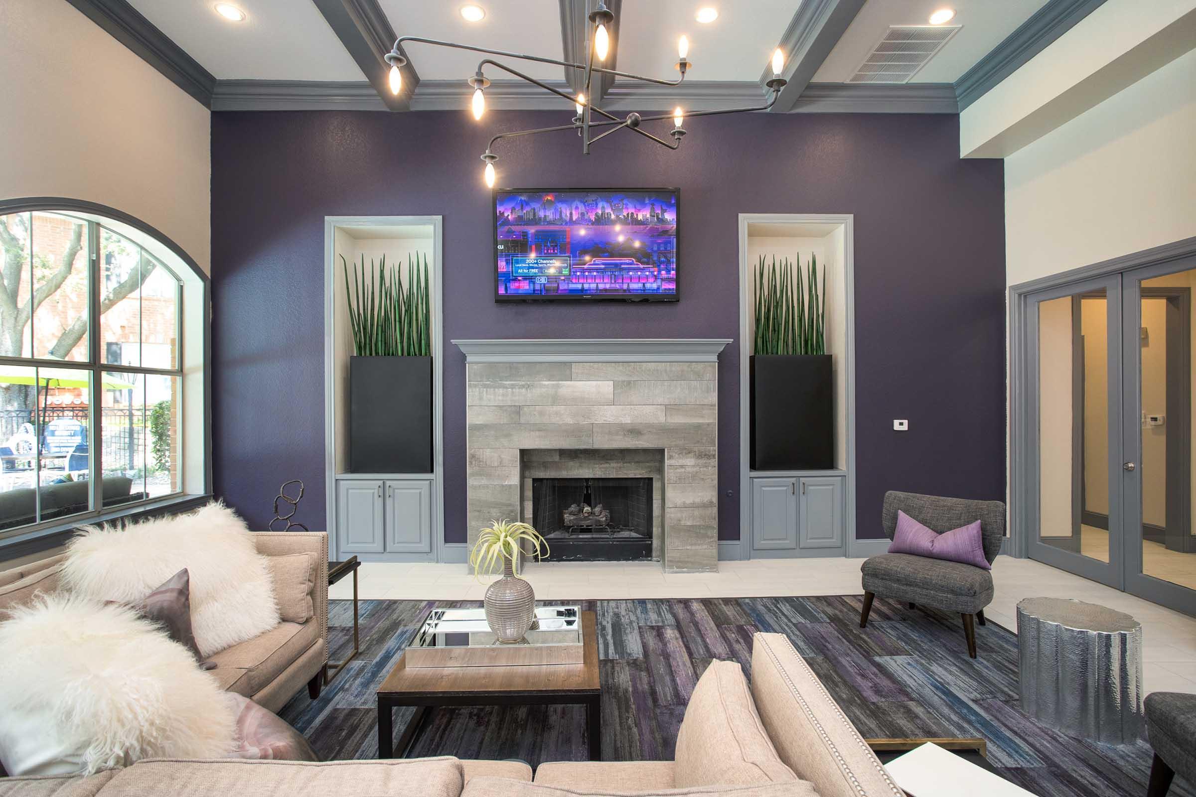 A modern living room featuring a light-colored sofa with decorative pillows, a gray accent chair, and a stylish coffee table. The walls are painted in a deep purple with two built-in shelves showcasing tall green plants. A fireplace adds warmth to the space, and a flat-screen TV is mounted above it. Bright overhead lights illuminate the area.