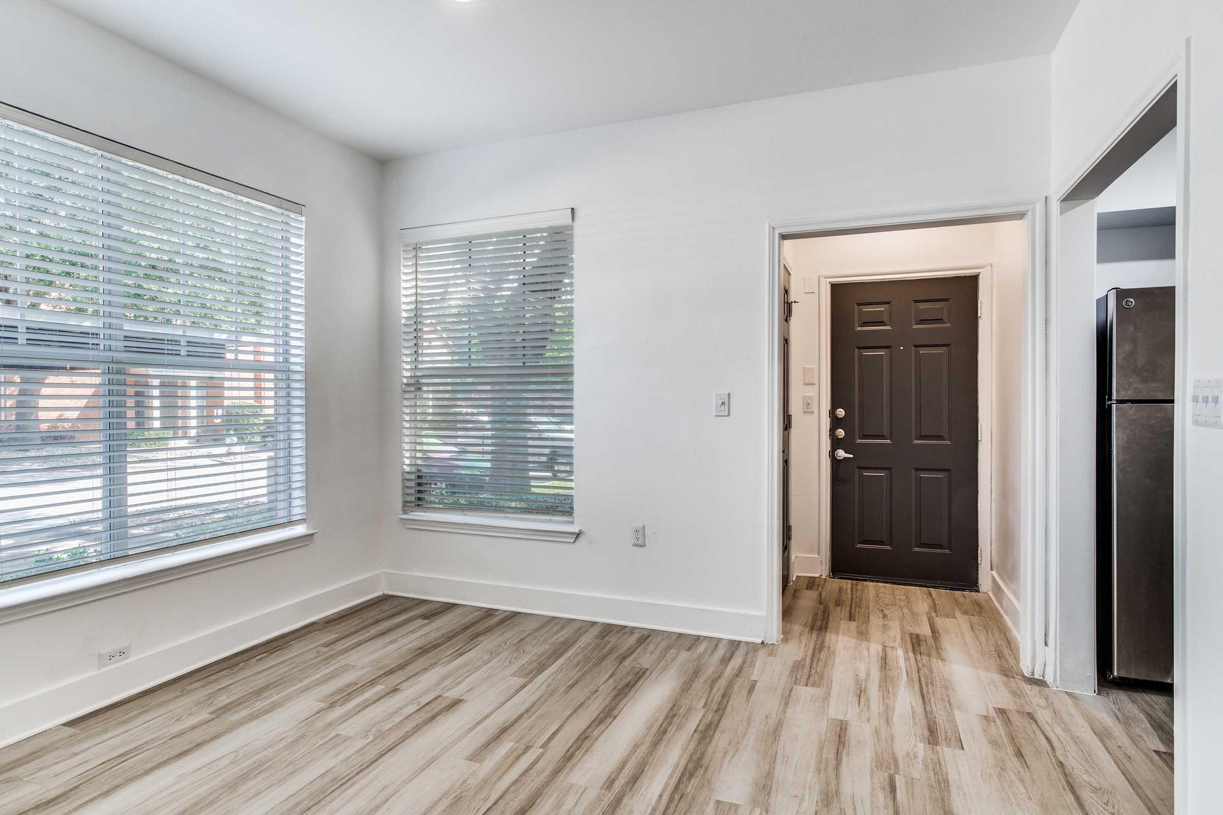 A well-lit interior space featuring large windows with blinds, a modern front door with a dark finish, and light wood-style flooring. The room has a clean and minimalist design, partially showing a nearby refrigerator in an adjacent area.