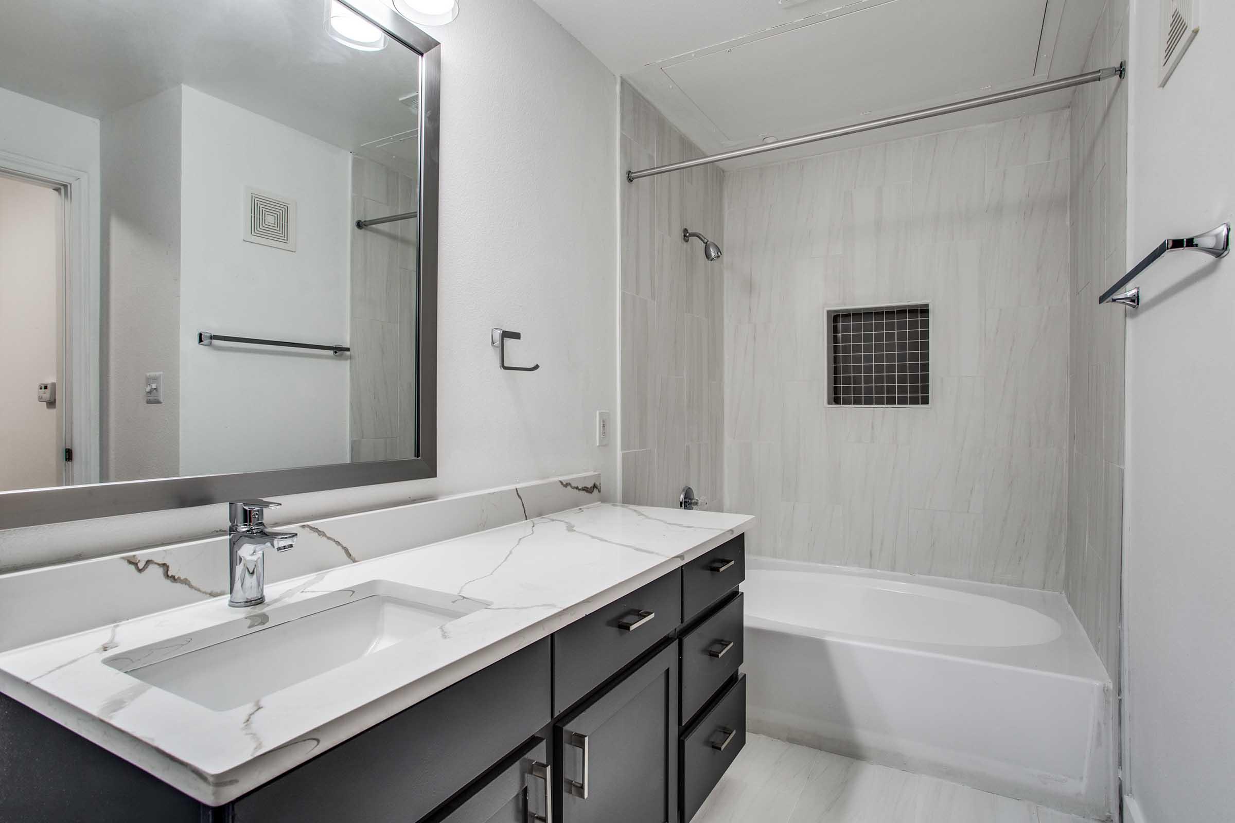 A modern bathroom featuring a white bathtub, a sleek black vanity with a marble countertop, and a large mirror. The walls are tiled in light colors, and there is a glass shower partition. Soft lighting enhances the clean and contemporary design.