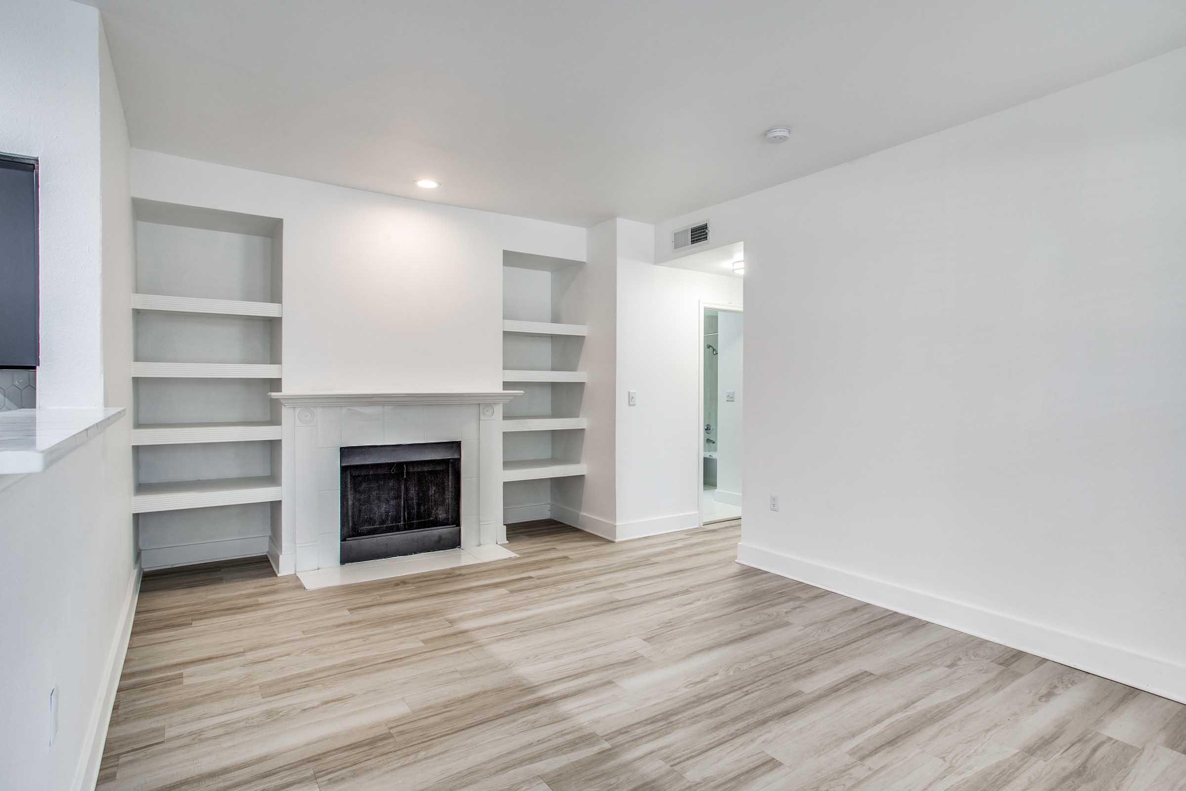 An empty room with light-colored walls and hardwood flooring. It features a white fireplace with a simple mantle, flanked by built-in shelving units. The room is well-lit with natural light and has a doorway visible at the back leading to another space.