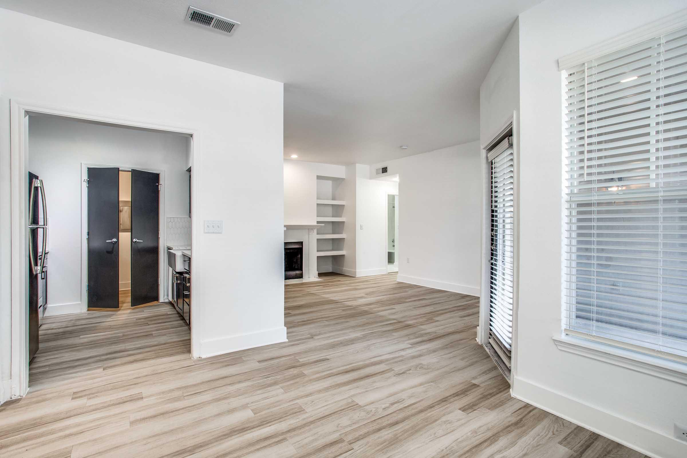 Spacious, modern interior of an apartment featuring light-colored wood flooring, white walls, and large windows providing natural light. The layout includes a living area with a fireplace, an open view towards the kitchen area, and a sliding door leading outside.
