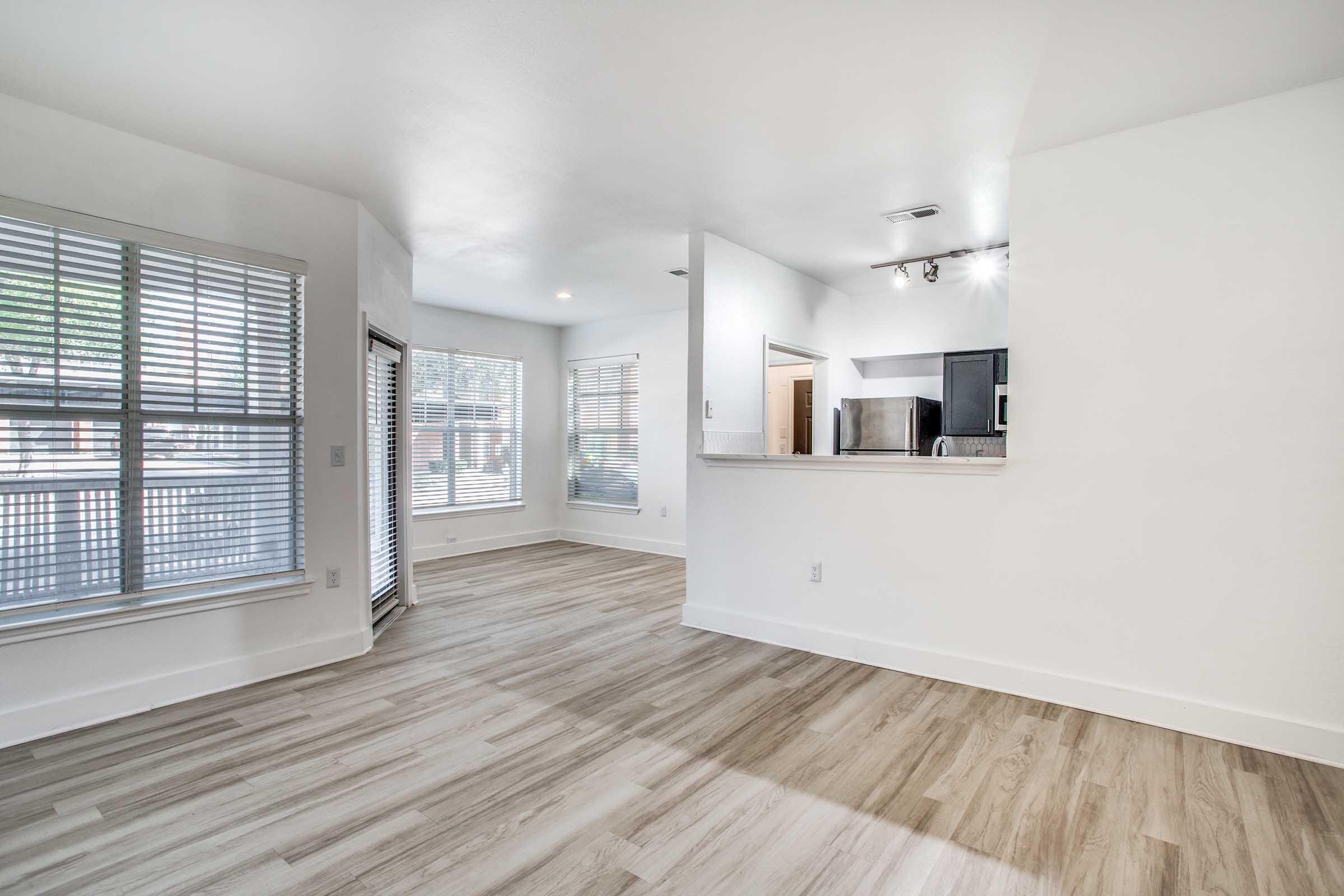 Spacious, modern living area featuring light-colored walls, large windows with blinds, and a seamless transition from the main room to an open kitchen. The floor is adorned with light wood-like planks, creating a warm and inviting atmosphere. Natural light brightens the space.