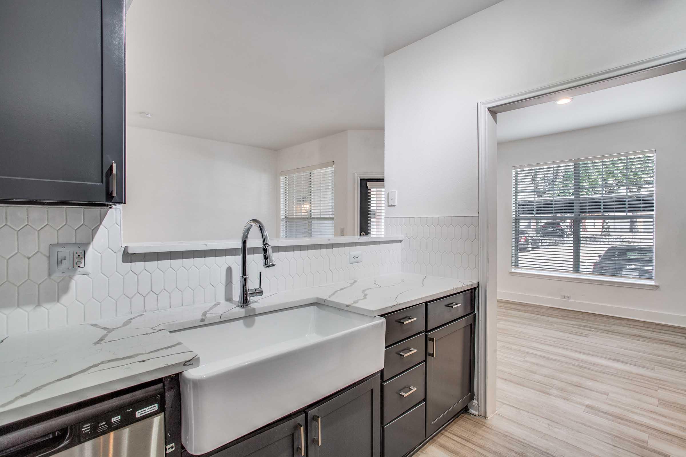 Modern kitchen featuring a farmhouse sink, dark cabinetry, and white hexagonal backsplash. The counter has a marble design, and a window allows natural light to fill the space. A doorway leads to a well-lit living area with large windows and wooden flooring.