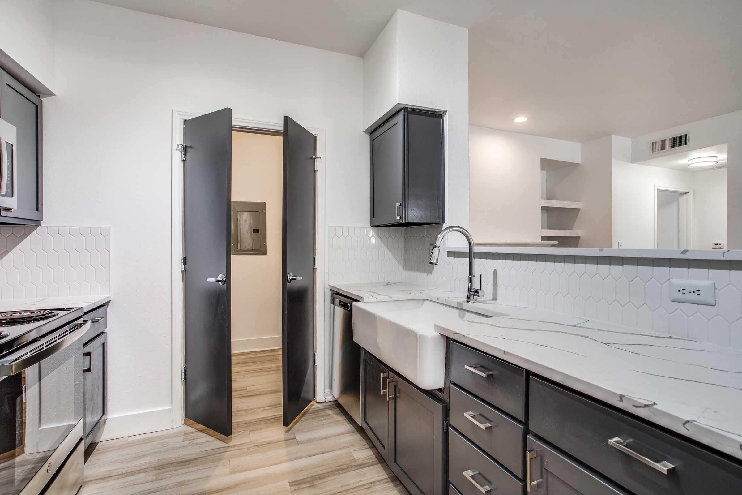 A modern kitchen featuring dark cabinetry and a white farmhouse sink. The countertops have a marble design, and there is a doorway leading to another room. The floor is made of light-colored wood. The space is well-lit, showcasing a clean and contemporary design.