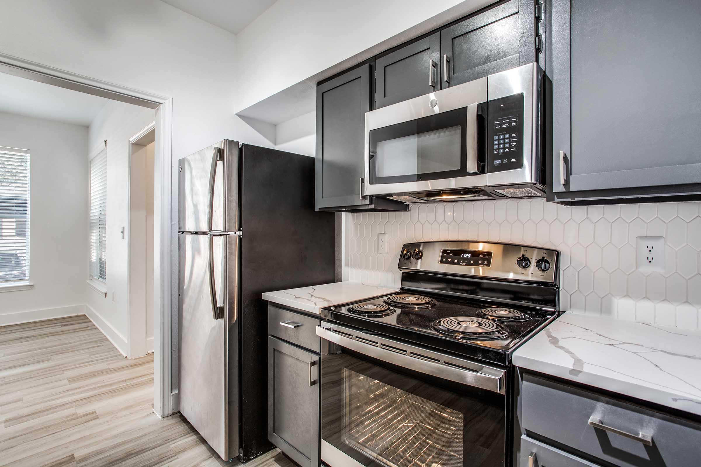 Modern kitchen featuring dark cabinets, stainless steel appliances including a refrigerator, microwave, and oven. A sleek stove with burners and a countertop with a white marble design. Light wooden flooring and windows providing natural light to the space.