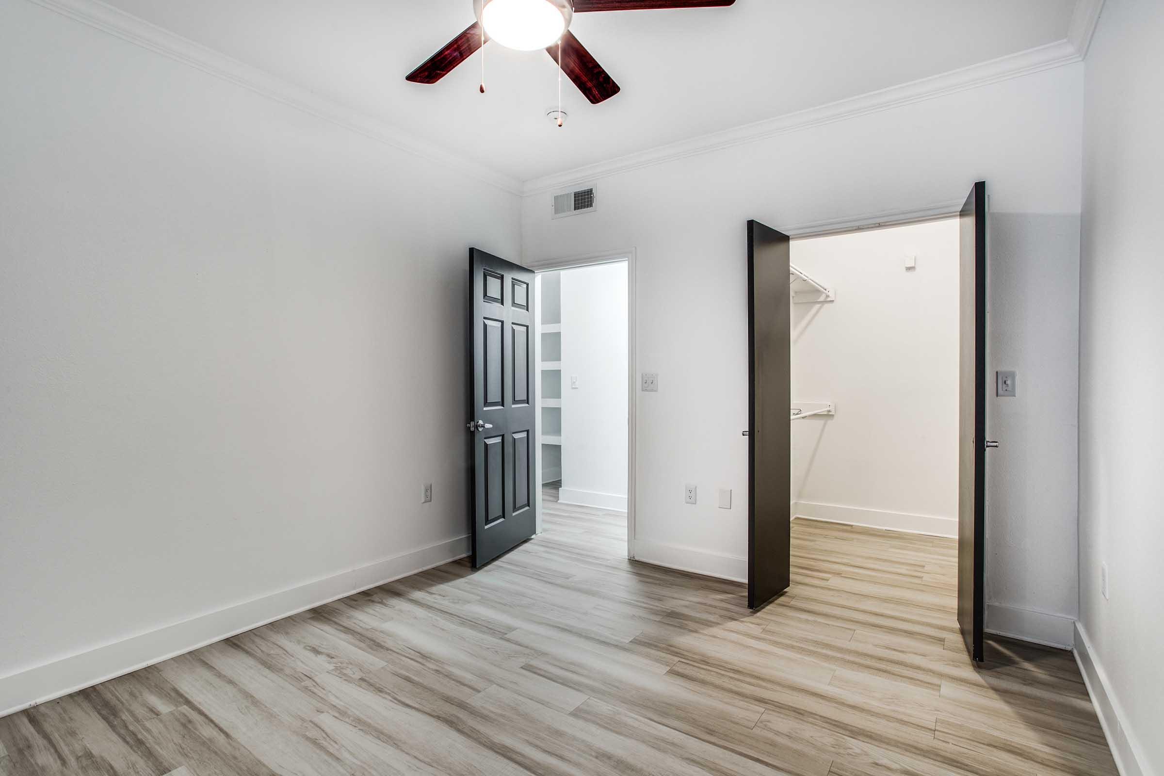 A well-lit interior room with light wood flooring, featuring two open doors leading to a closet space. The walls are painted white, and there is a ceiling fan with dark wooden blades. The room has a modern design, providing a sense of spaciousness and simplicity.