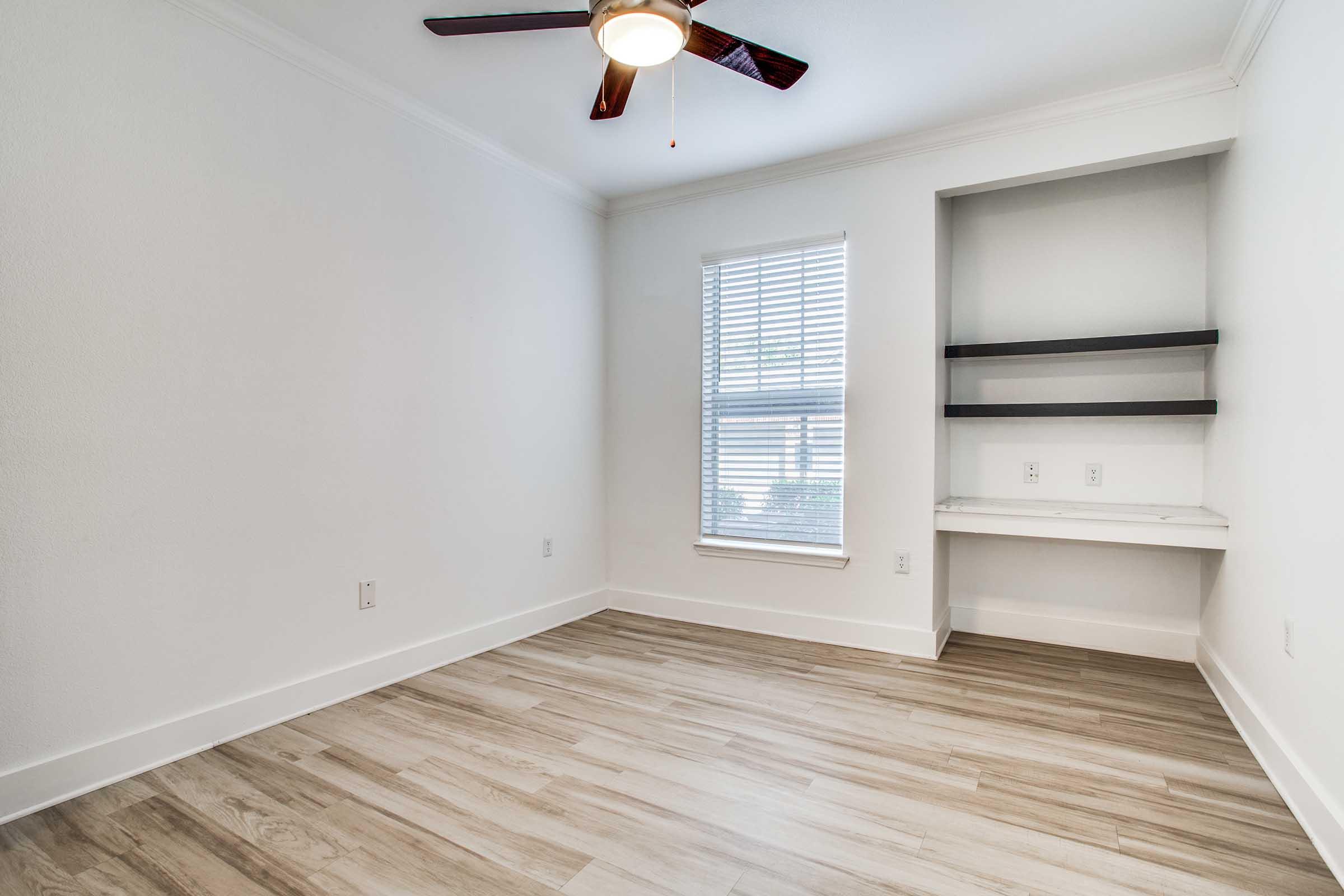 A vacant room with light-colored walls and wood-look flooring, featuring a ceiling fan with wooden blades. There is a window with blinds allowing natural light to enter, and a small built-in shelf unit against one wall. The overall atmosphere is modern and minimalistic.