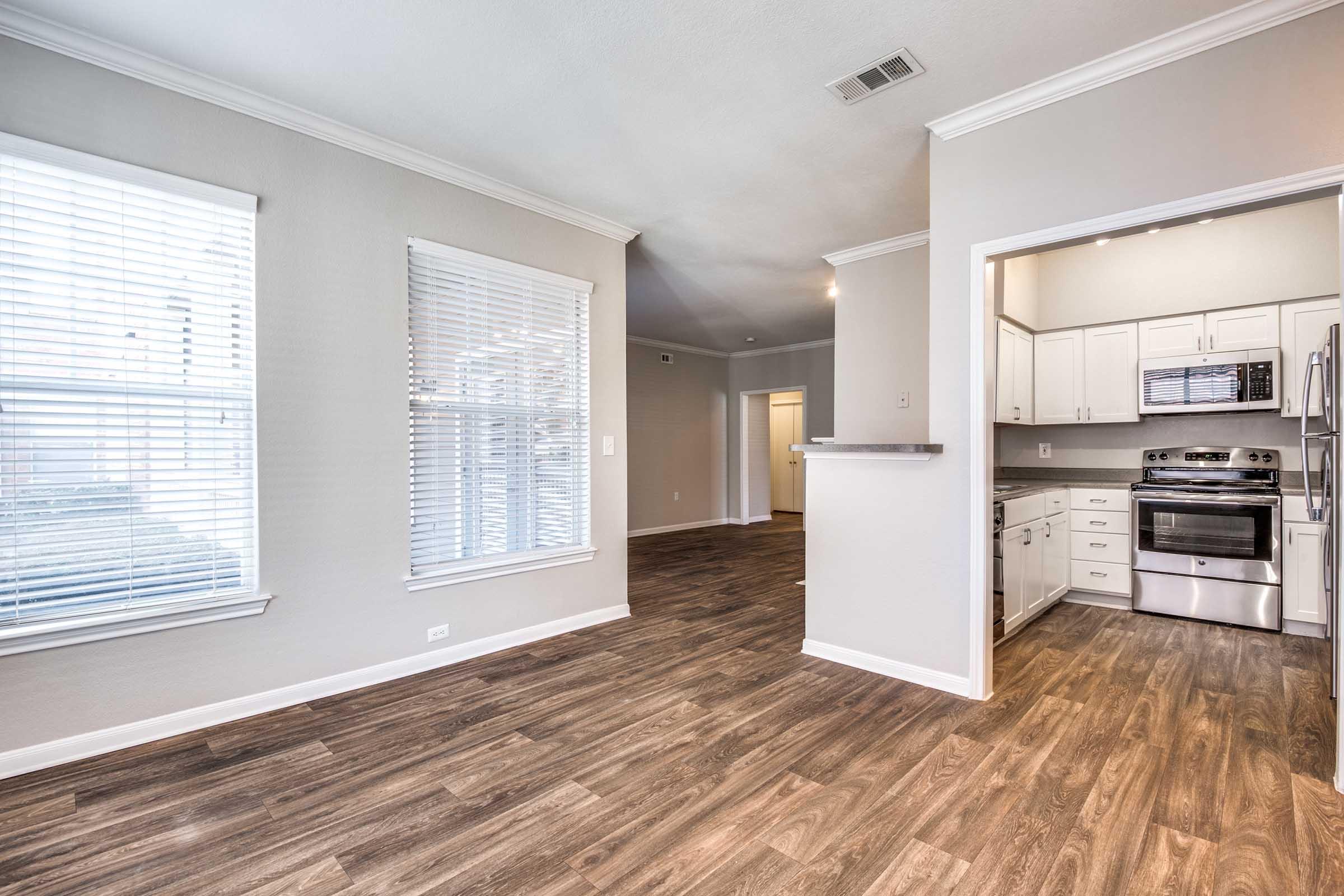Spacious living area with large windows letting in natural light, featuring light gray walls and wood-style flooring. An open kitchen visible in the background, equipped with stainless steel appliances and white cabinetry. The layout promotes a bright and inviting atmosphere, ideal for comfortable living.