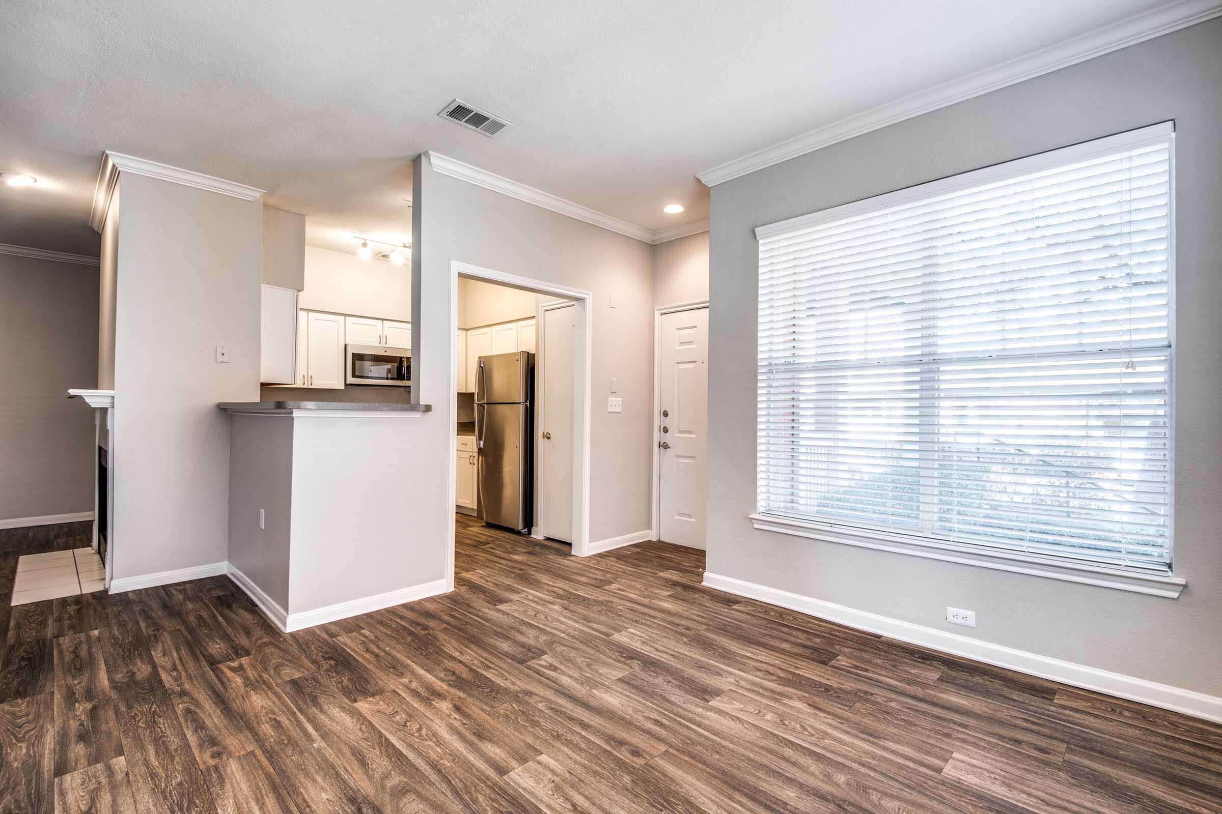 A bright, modern living space featuring hardwood flooring, neutral walls, and a large window with blinds. The open layout includes a kitchen visible in the background, with white cabinetry and stainless steel appliances. The room has a welcoming atmosphere, ideal for relaxation or entertaining.