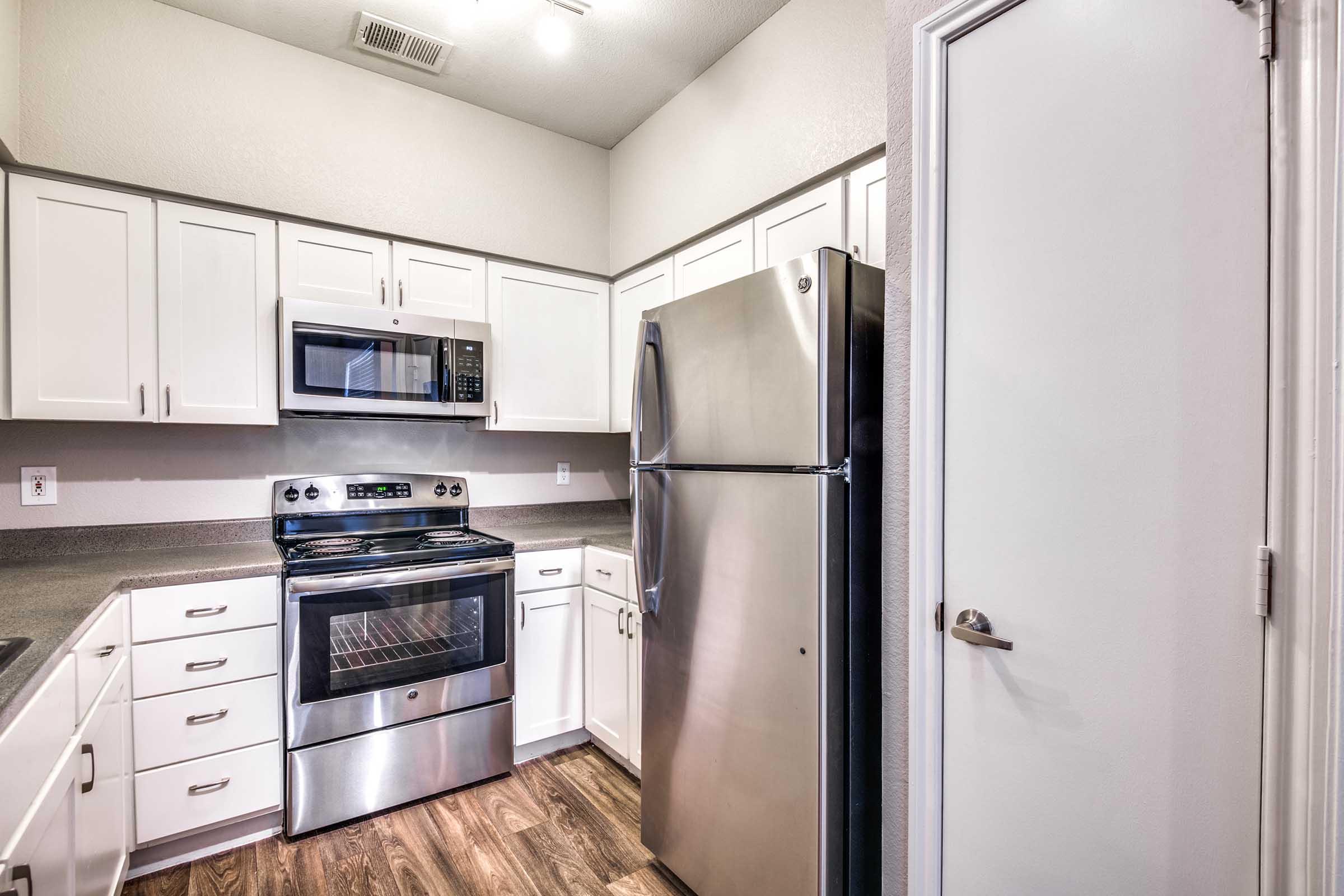 A modern kitchen featuring white cabinetry, stainless steel appliances including an oven, microwave, and refrigerator, along with a countertop. The flooring is wood-like laminate, and there is a door leading to another room. Natural light illuminates the space, enhancing its clean and contemporary design.