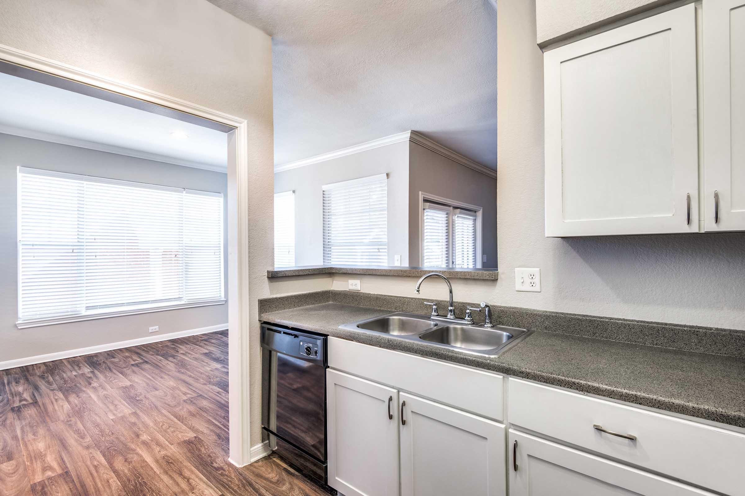 Modern kitchen with a double sink and dishwasher, featuring gray countertops and white cabinetry. The background shows a well-lit living area with large windows and hardwood-style flooring.
