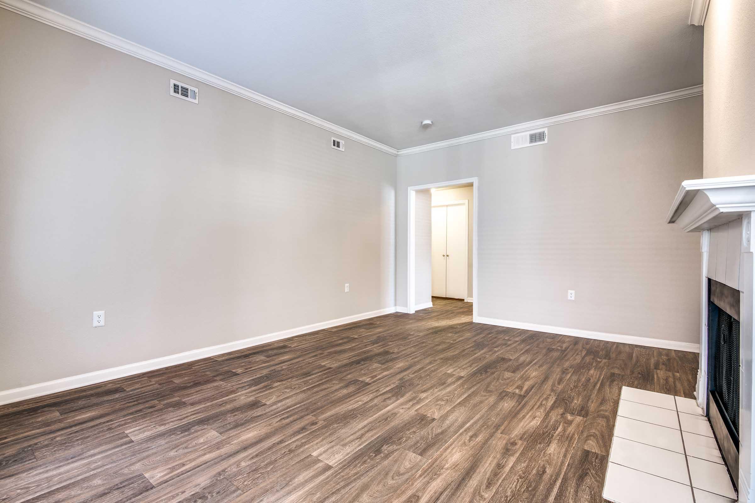 Empty living room with light-colored walls and a wood-look floor. There is a fireplace with a white mantel on the right. A doorway leads to another room on the left. The space is well-lit and open, creating a spacious and inviting atmosphere.