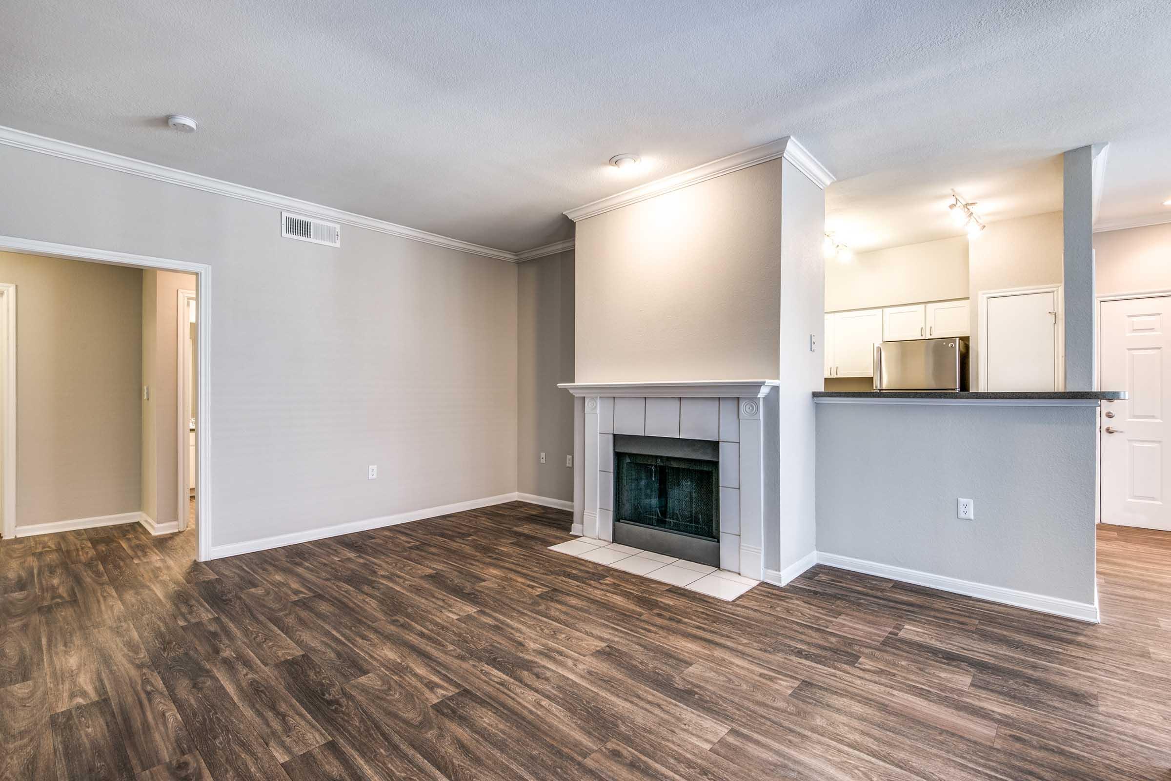 A spacious living area featuring a fireplace, hardwood-style flooring, and neutral-colored walls. To the left, an open doorway leads to another room. The kitchen area is visible in the background, showcasing modern appliances and soft lighting. Bright and inviting atmosphere suitable for comfortable living.