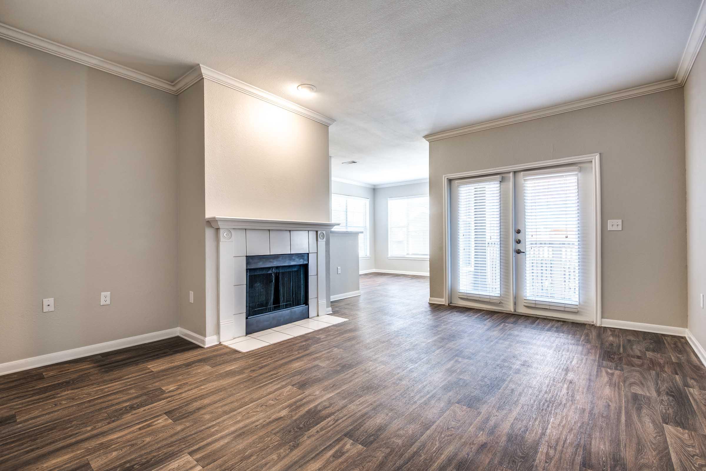 Spacious living room with hardwood-style flooring, a fireplace with a white mantel, and large windows. A glass door leads to a balcony, allowing natural light to fill the room. The walls are painted in a light, neutral color, creating a cozy atmosphere.