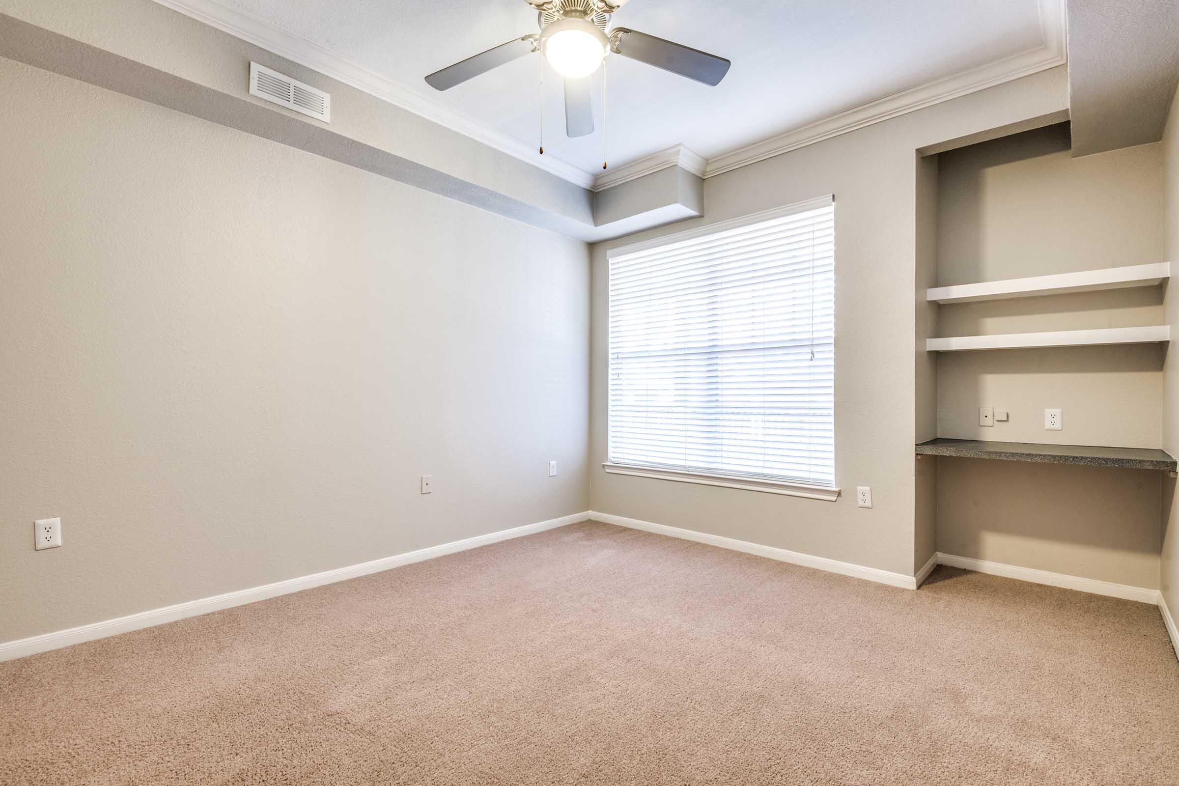 A vacant room with light gray walls and plush beige carpeting. A window with blinds lets in natural light, and there is a ceiling fan overhead. A small built-in shelf is visible in one corner, and the room appears spacious and well-lit, ideal for various uses.