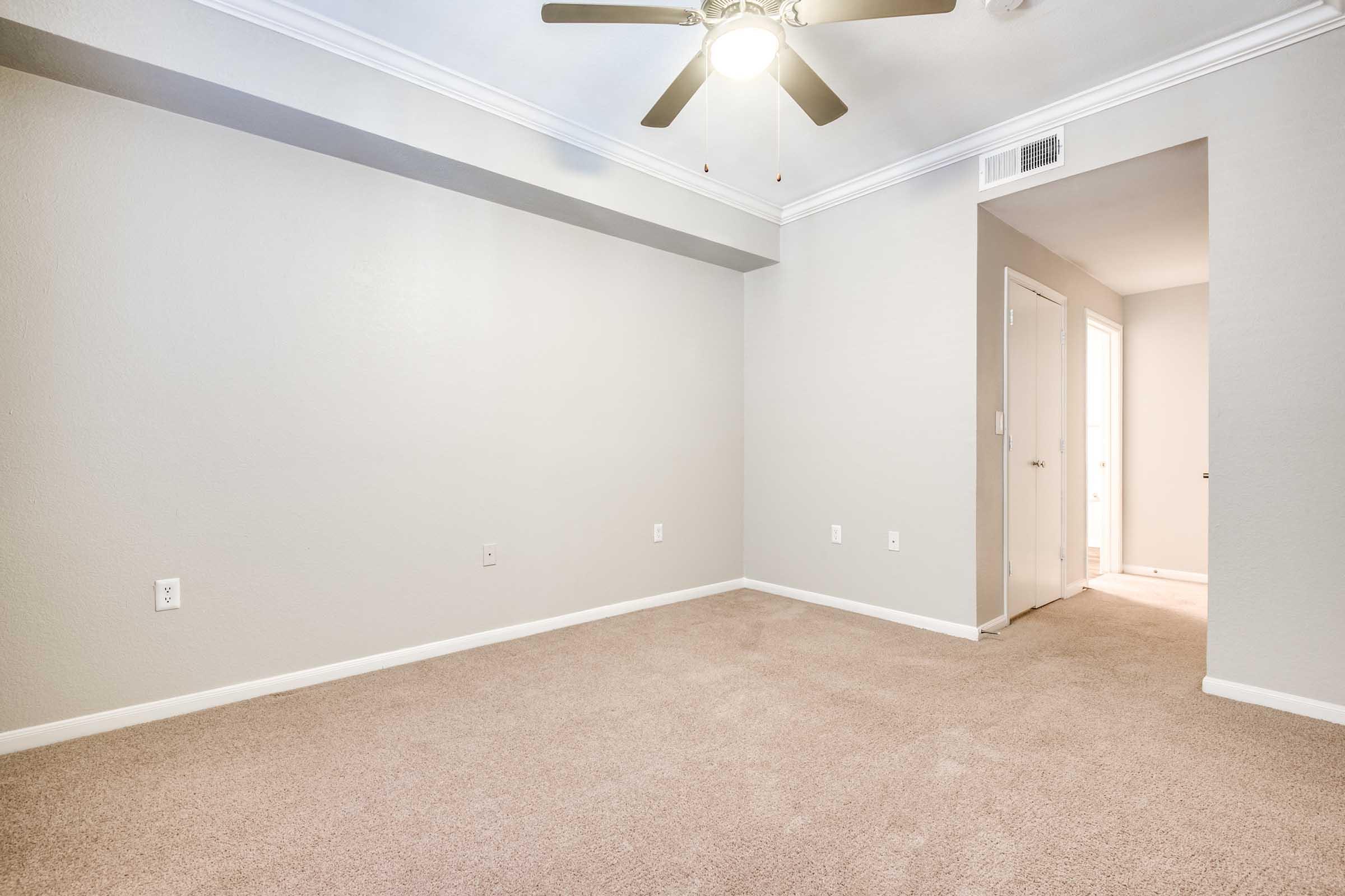 A spacious, empty room featuring light gray walls and beige carpet. A ceiling fan is mounted in the center, providing ample lighting. There is an open doorway on the right leading to a hallway with a glimpse of a bathroom or another room. The overall atmosphere is clean and ready for furnishing.
