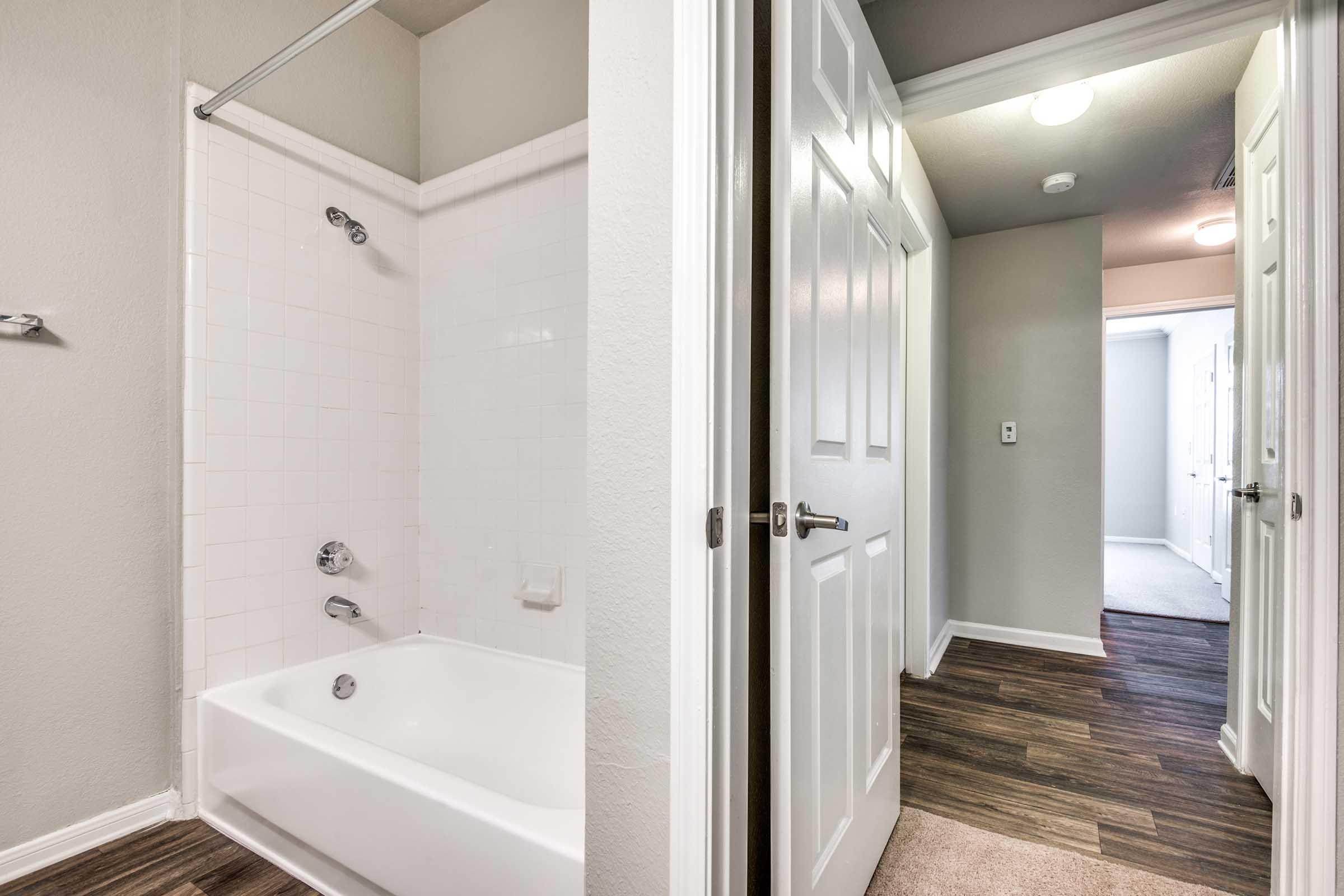 View of a modern bathroom with a shower and bathtub on the left, featuring white tiles and a chrome showerhead. On the right, a hallway leads to additional doors and rooms, showcasing wood-style flooring and neutral-colored walls. The lighting is soft and inviting.