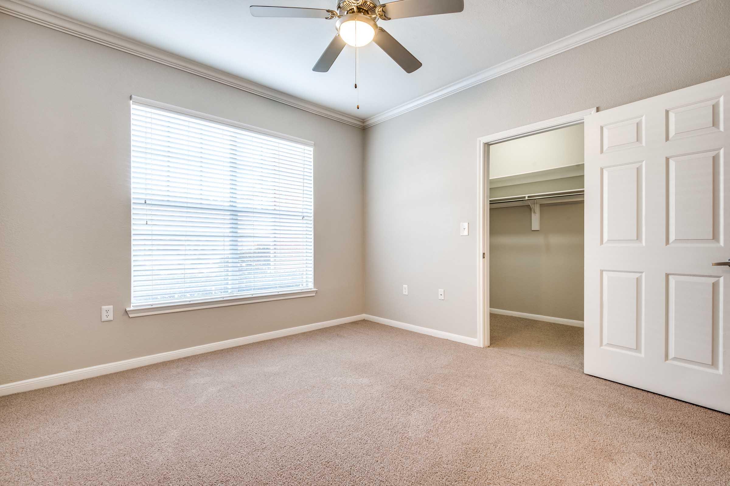 A bright, empty bedroom featuring light beige walls and carpet, with a ceiling fan and a window covered by white blinds. A closet is visible through an open door, and the overall space appears clean and well-maintained, ready for new furnishings.