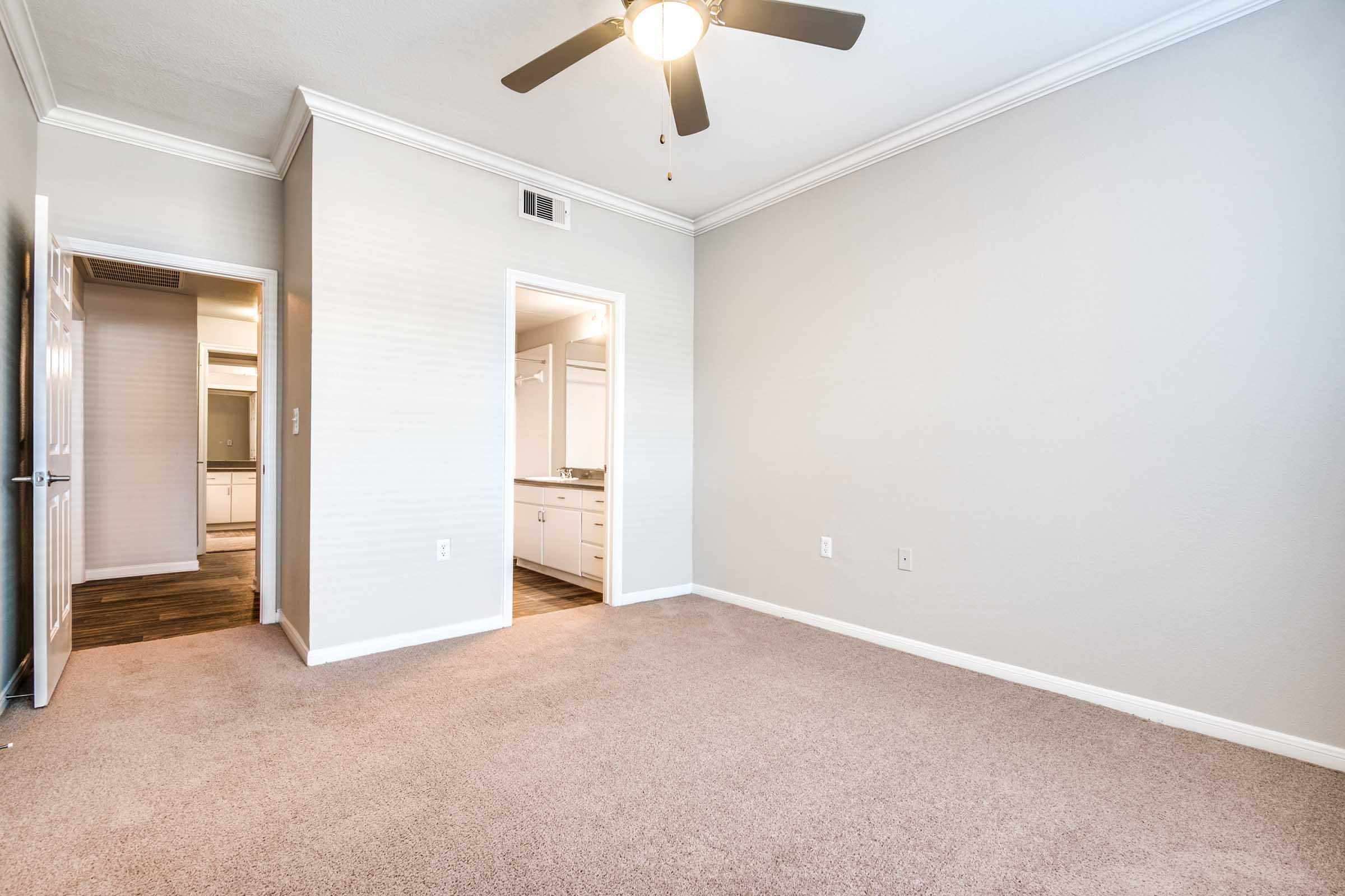 A spacious, empty room with light-colored walls and soft beige carpet. A ceiling fan is visible, and there are doorways leading to other areas. The room has a bright, airy feel, with natural light coming through the openings.