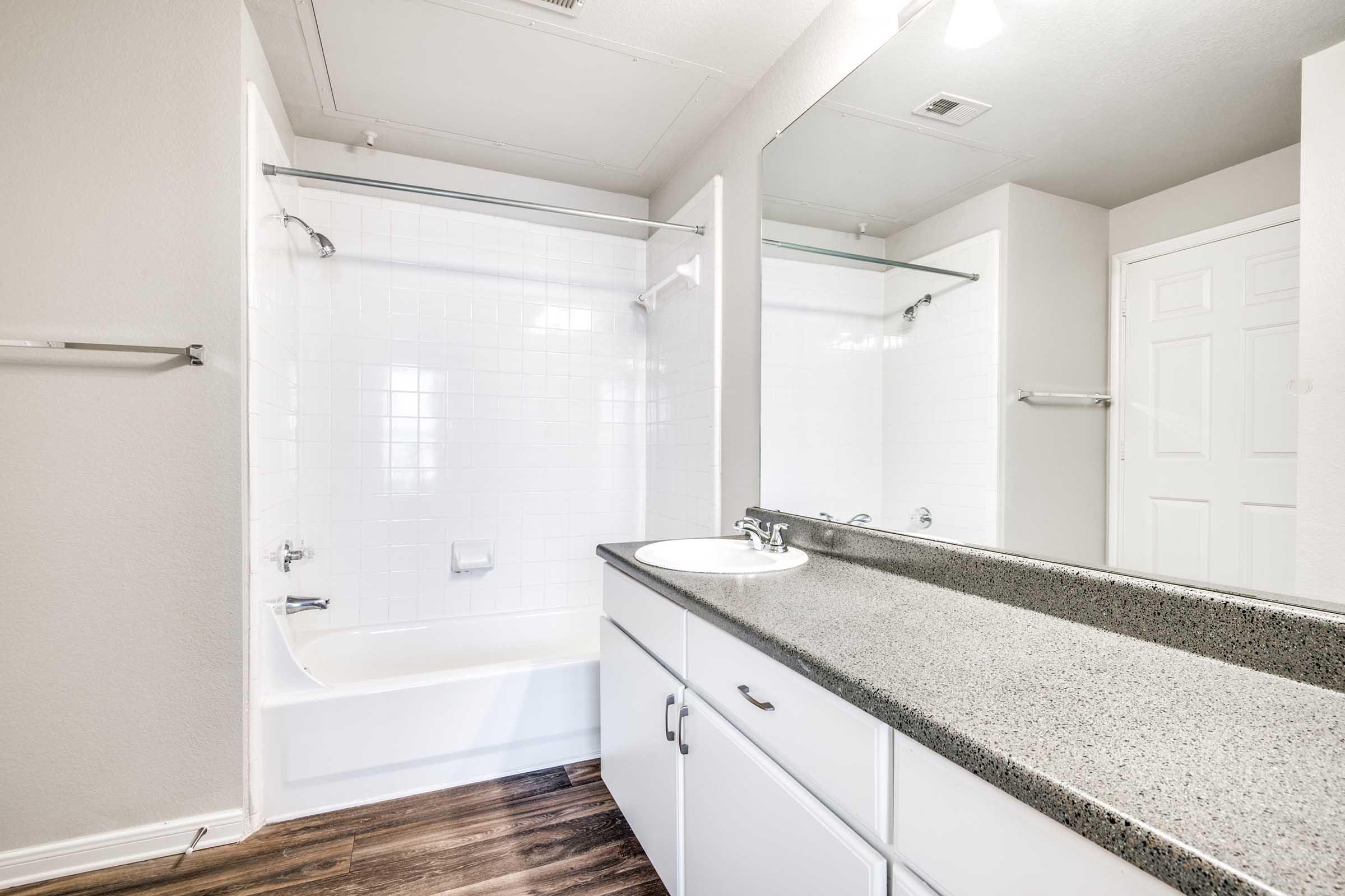 A modern bathroom featuring a shower/tub combo, a single sink with a countertop, and a large mirror. The walls are painted in neutral tones, and the floor has a wood-like appearance. The space is well-lit by natural light coming through a window.