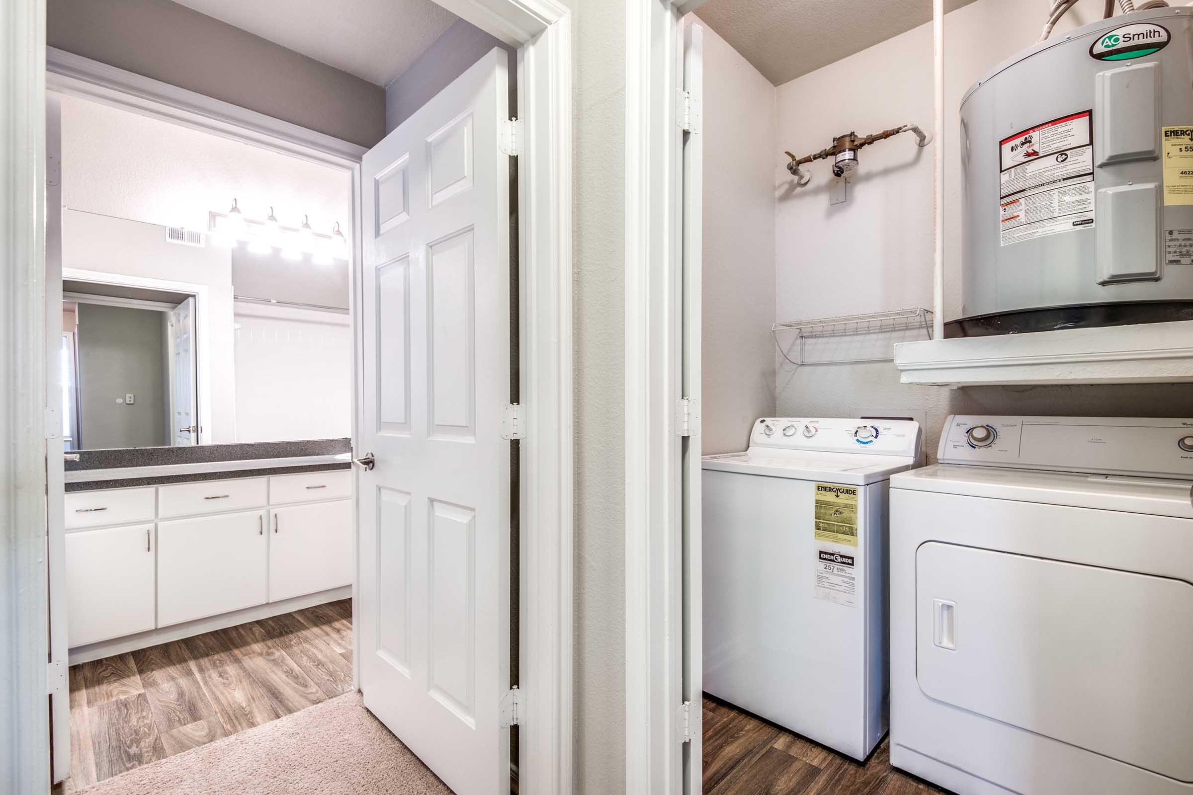 A laundry area featuring a washing machine and dryer, with a water heater mounted on the wall. The space includes doors leading to a bathroom and another room, with white cabinetry and light-colored walls. Natural light illuminates the area, highlighting the wood-style flooring.