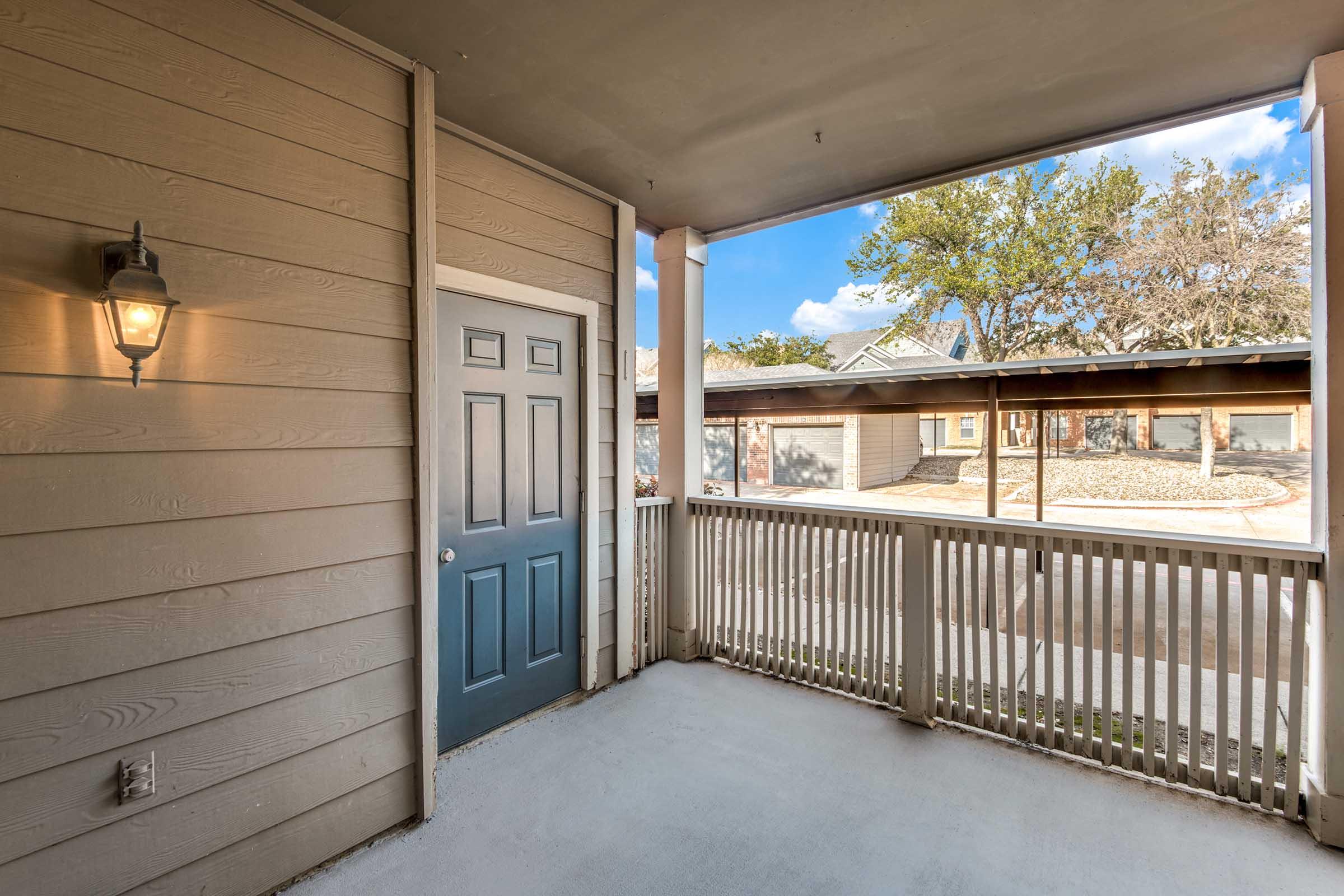 A covered porch area featuring a blue door, wooden siding, and a lantern on the wall. The porch is spacious and includes a railing, with a view of trees and nearby houses in the background under a clear blue sky.