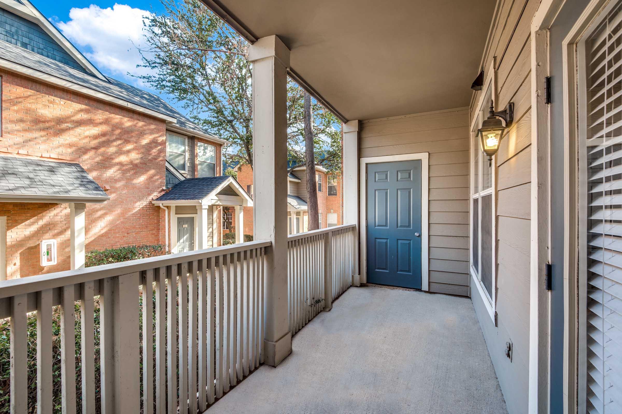 A view of a covered porch with a light fixture on the wall, featuring a blue door and a railing. The surroundings include brick buildings and trees, with sunlight filtering through. The porch is spacious and neatly maintained, providing a welcoming entrance to the home.