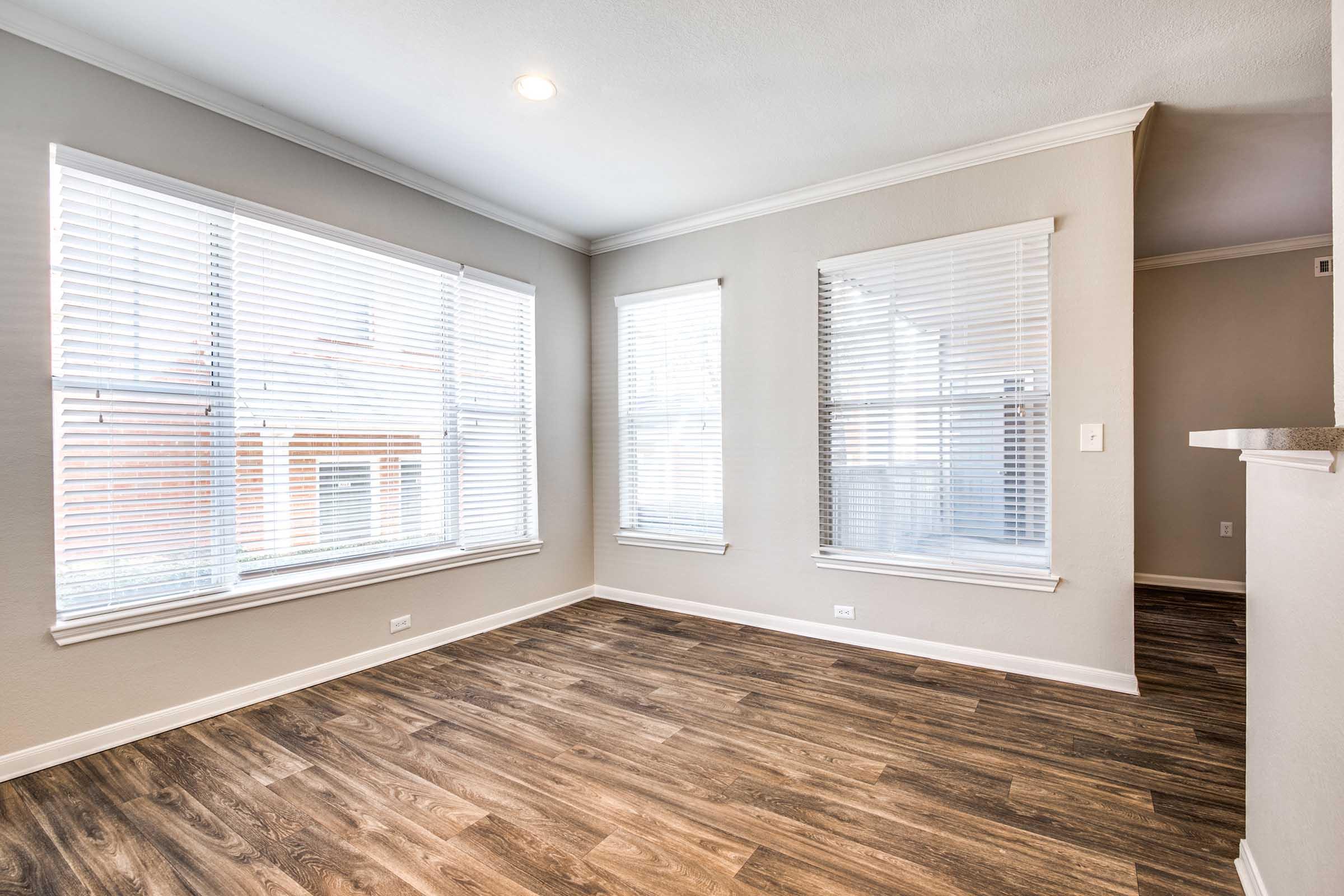 A bright, empty living room featuring large windows with white blinds, allowing natural light to flood in. The walls are painted in a light neutral color, and the floor is covered with wood-like laminate. The room has a clean and modern appearance, suitable for various decor styles.