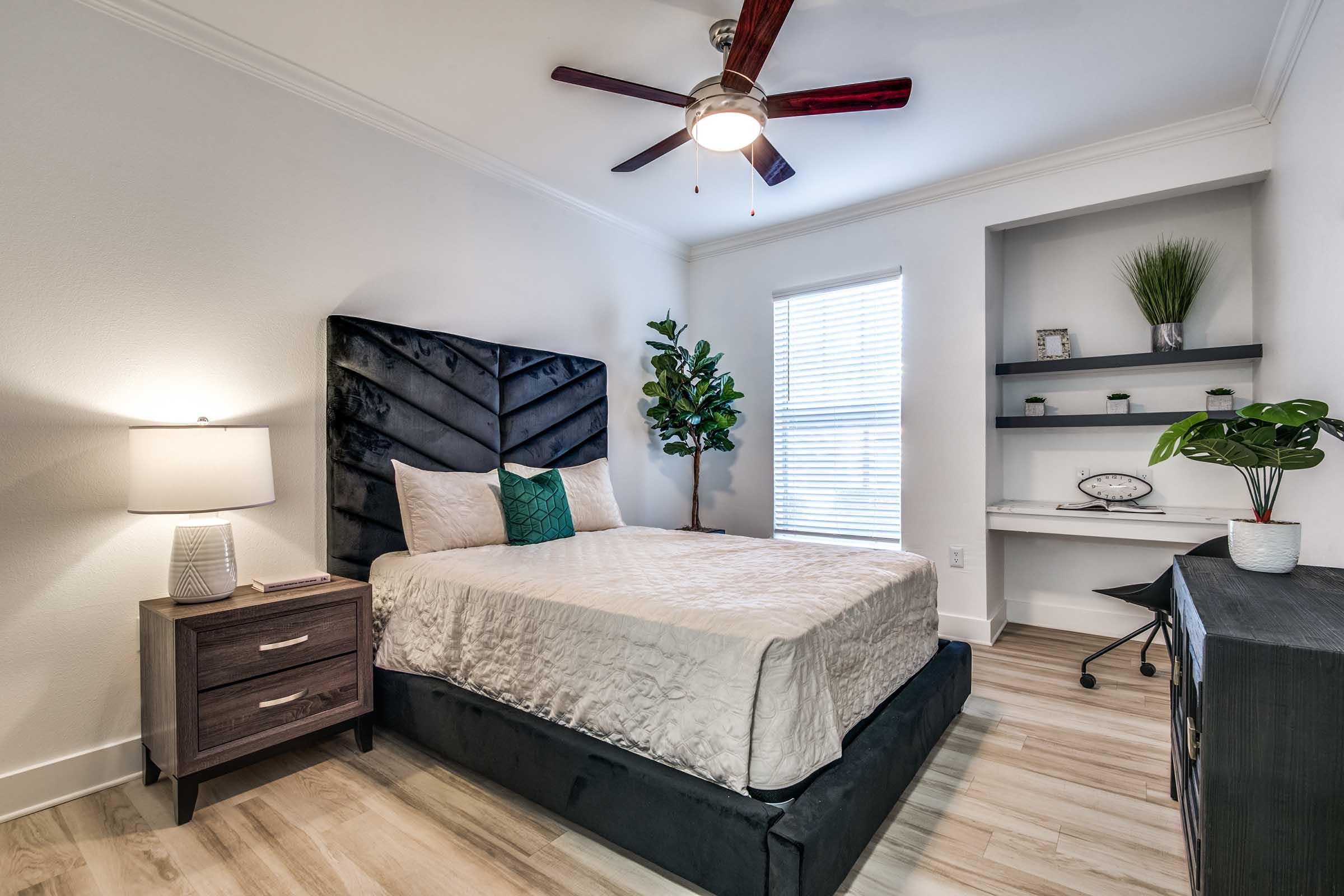 Modern bedroom featuring a dark upholstered bed with decorative pillows, a bedside table with a lamp, a potted plant, and a small desk with a chair. The room has light-colored walls, wooden flooring, and a ceiling fan. A window allows natural light to brighten the space.