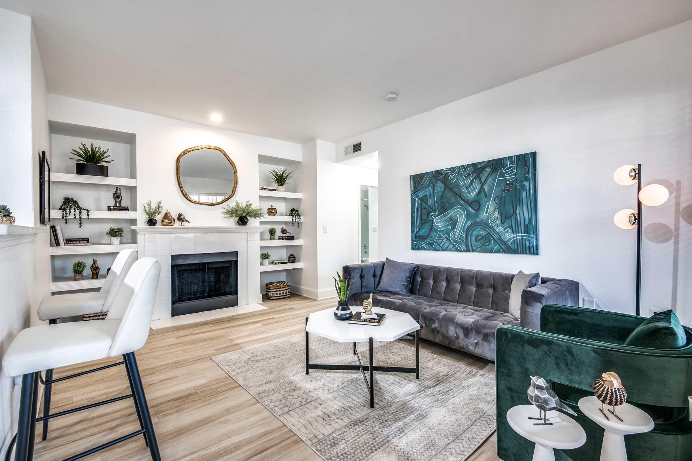 A modern living room featuring a gray sofa, green accent chair, and a coffee table. There is a fireplace with a white mantel, shelves decorated with plants and decorative items, and a large round mirror above the fireplace. The walls are painted white, and the flooring is light wood.
