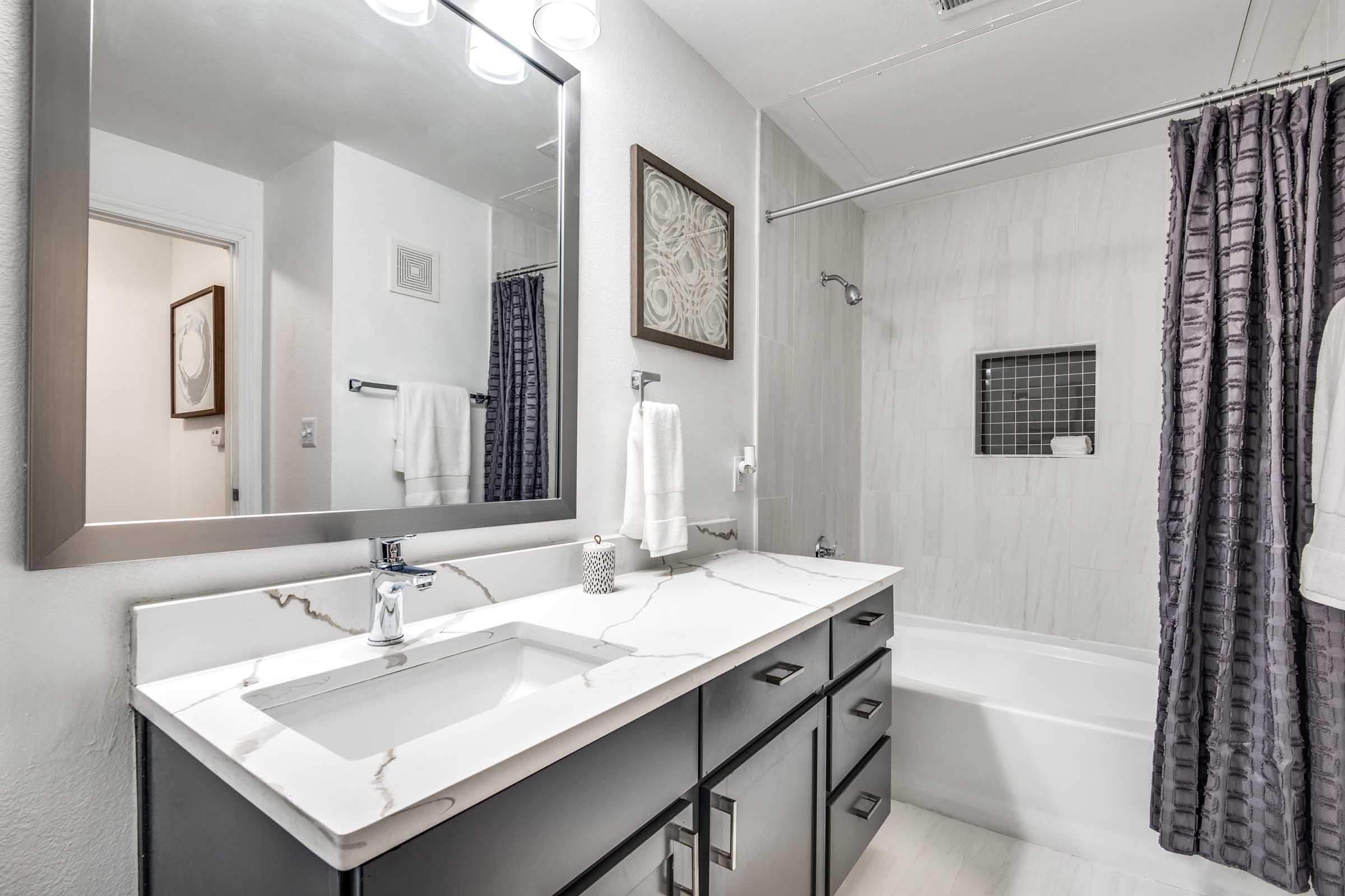 A modern bathroom featuring a sleek vanity with a white and gray marble countertop, a rectangular mirror, and a bathtub with a dark gray shower curtain. The walls are light-colored, and there are decorative elements like a framed artwork and neatly arranged towels.