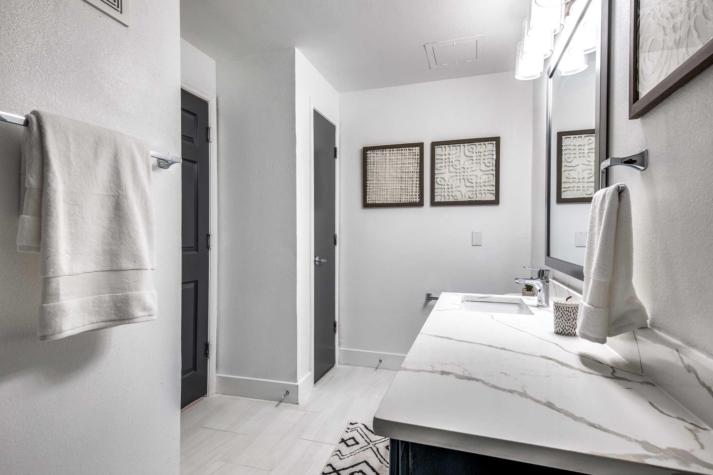 A modern bathroom featuring a white marble countertop with a sink, framed artwork on the walls, a towel rack with a white towel, and a grey door leading to another space. The walls are painted white, and the floor has light-colored tiles, creating a clean and contemporary aesthetic.