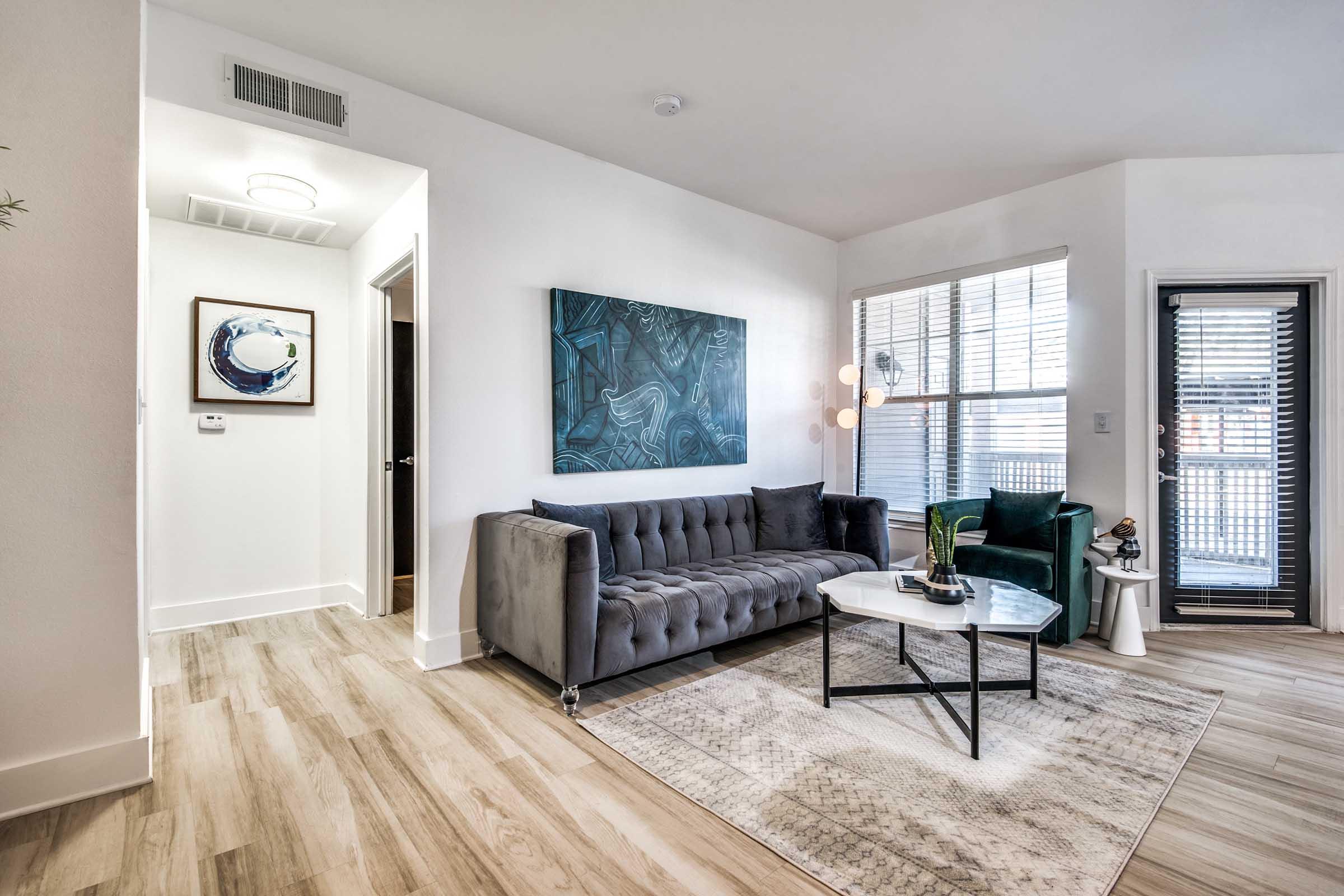 A modern living room featuring a gray tufted sofa and a round coffee table on a light area rug. There's an abstract blue artwork on the wall, a green armchair, and large windows with blinds letting in natural light. A doorway leads to another space, adding to the open feel of the room.