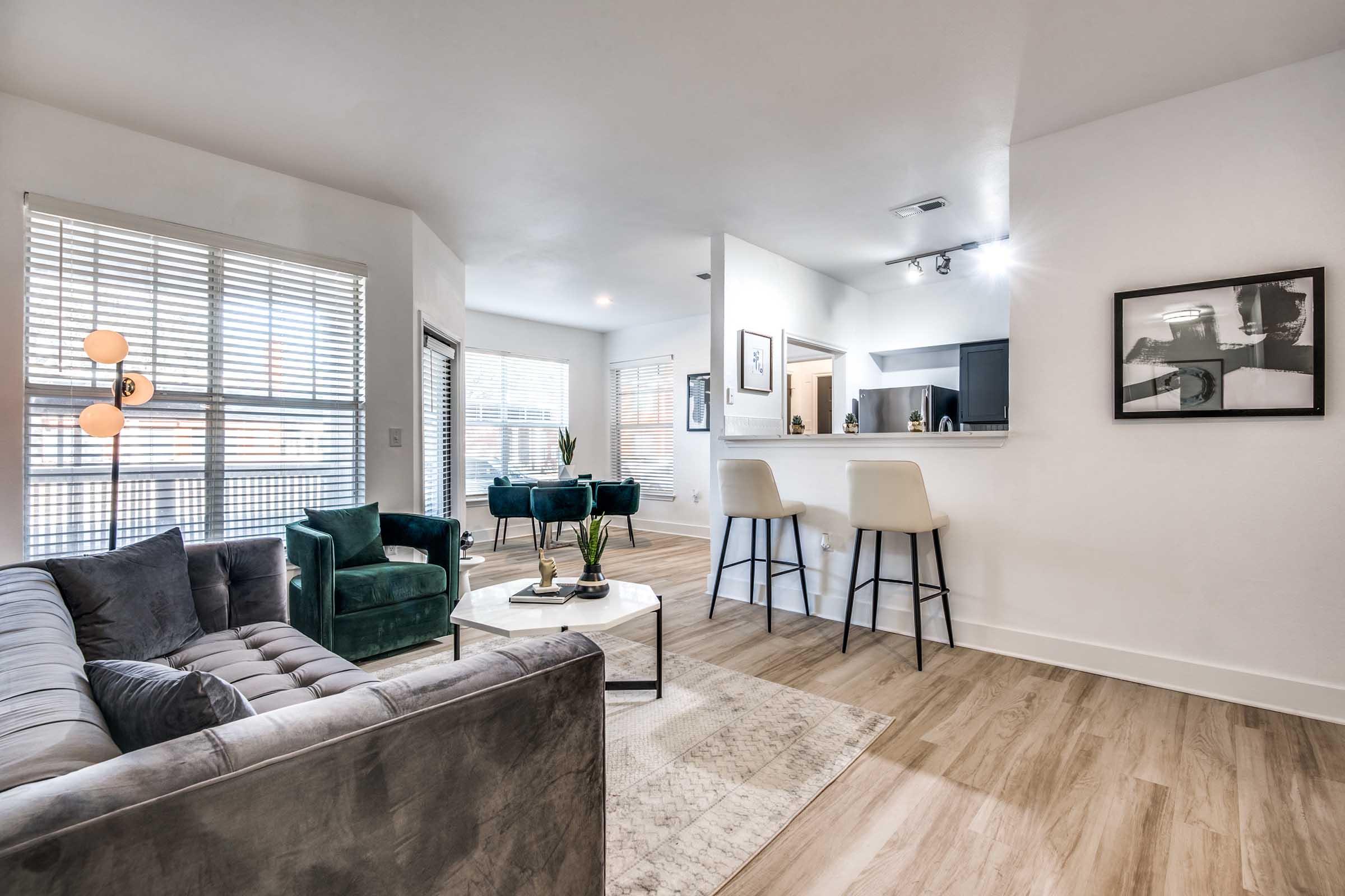A modern living space featuring a gray velvet sofa, a round coffee table, and two green chairs. A kitchen counter with bar stools is visible in the background, along with a dining area. Large windows let in natural light, and decorative elements include artwork and plants. The flooring is a light wood finish.