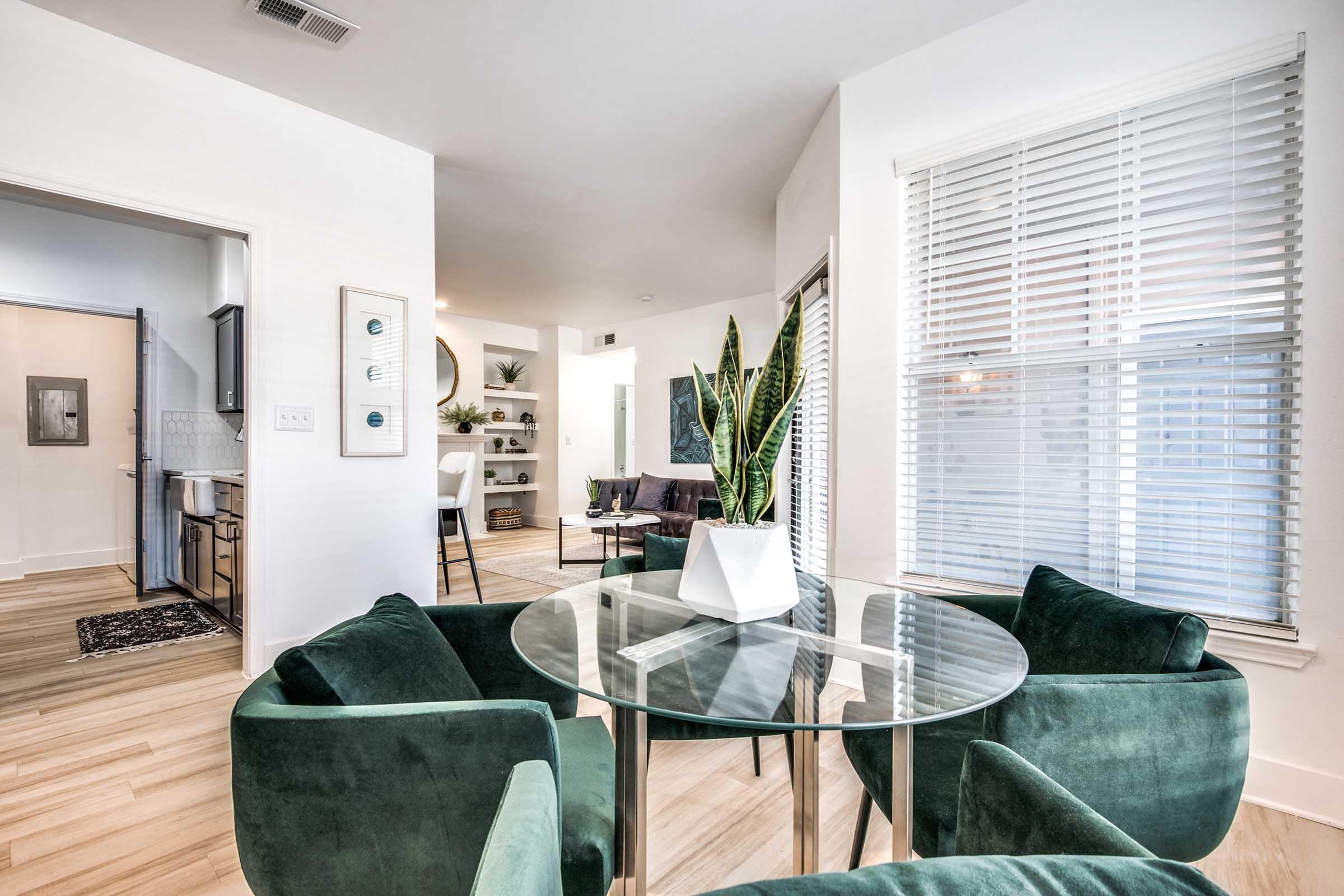 Modern interior of a bright living space featuring a glass dining table surrounded by green upholstered chairs. In the background, there's a stylish seating area with a plant, shelves with decor, and large windows allowing natural light. The decor is contemporary with a neutral color palette.