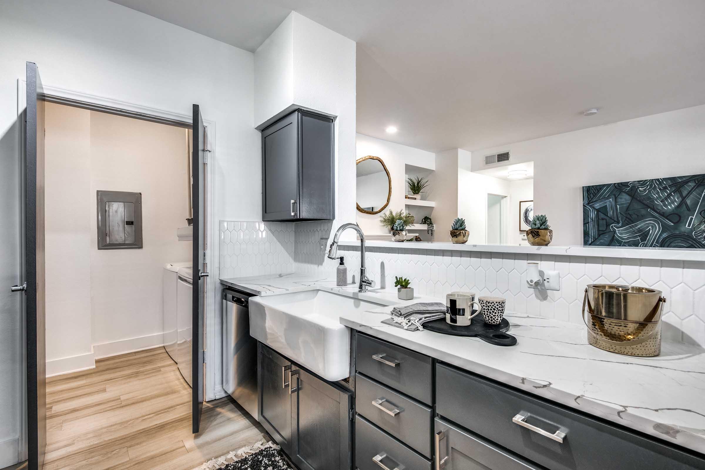 Modern kitchen featuring gray cabinets, white hexagonal tile backsplash, a large farmhouse sink, and marble countertop. A doorway leads to a laundry area, and decorative items like plants and a bowl are arranged on the counter. Natural light fills the space, enhancing the contemporary design.