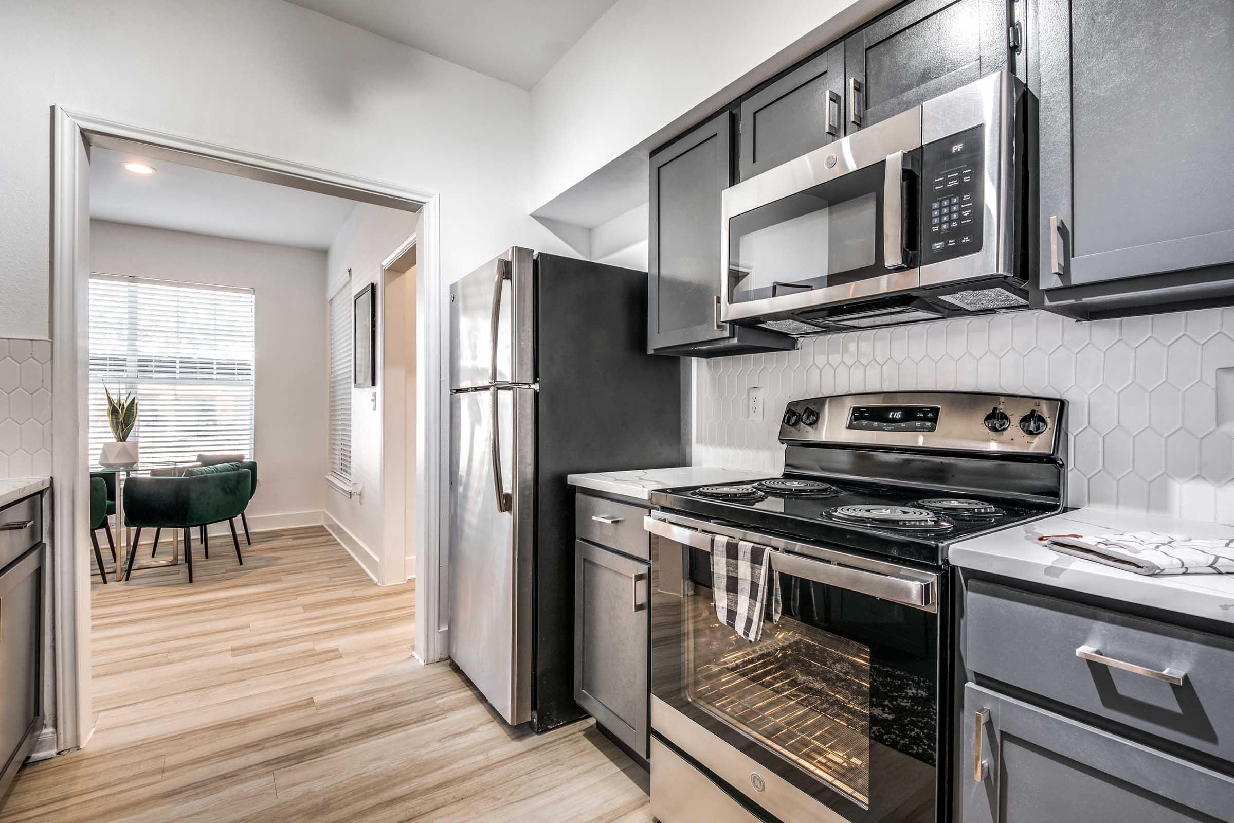 A modern kitchen featuring stainless steel appliances, including a microwave and oven, dark cabinets, and a light-colored backsplash. The kitchen is connected to a dining area with a small table and chairs, and there is natural light coming through a window. The flooring is a light wood finish.