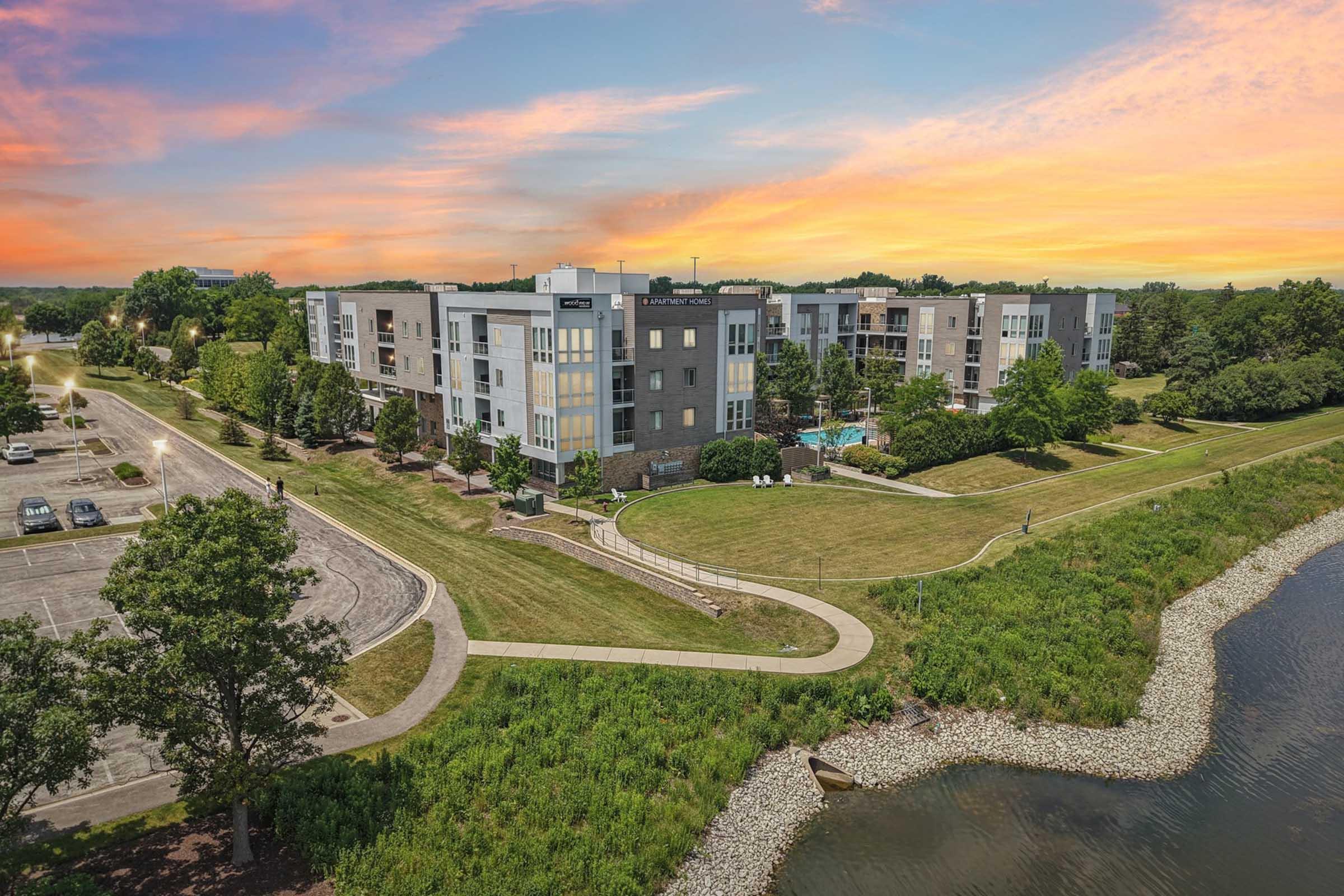 A modern apartment complex situated near a riverbank, surrounded by green lawns and trees. The evening sky features vibrant hues of orange and pink. A walking path meanders through the landscape, with a parking lot visible in the background. The scene conveys a serene residential environment.