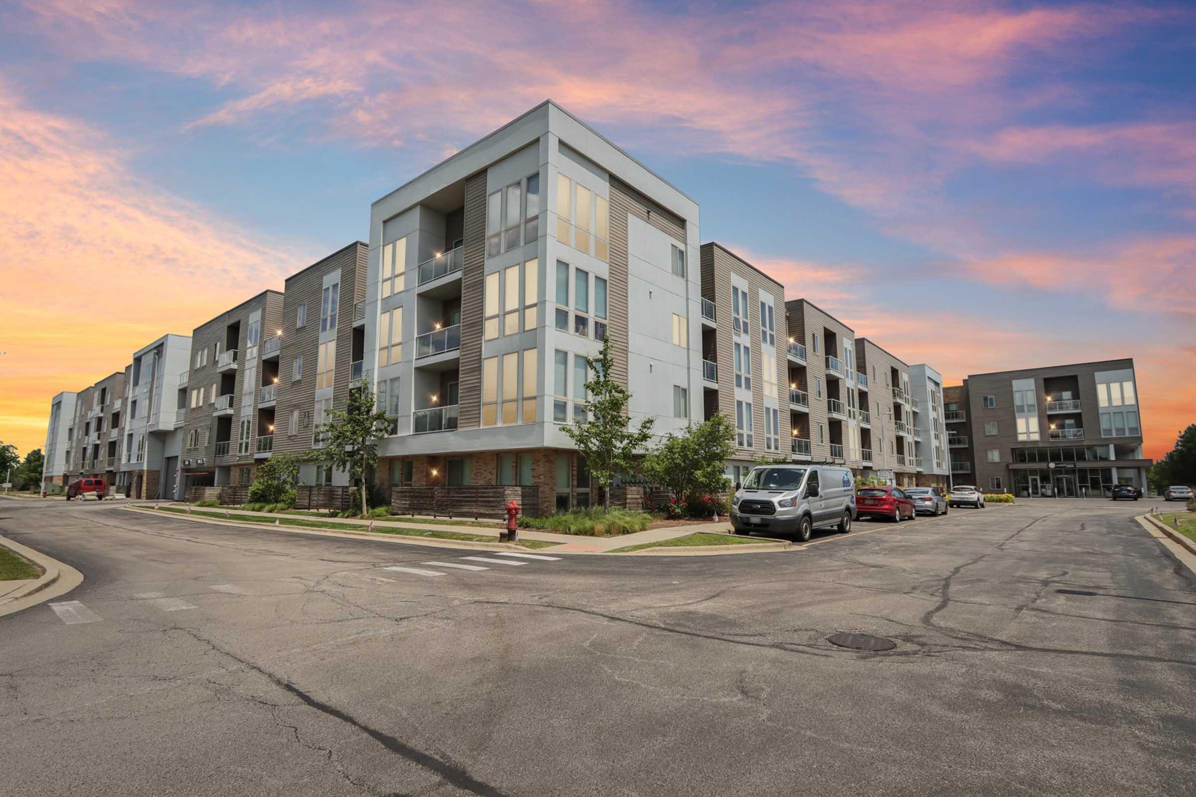 A modern apartment complex featuring multiple three-story buildings with large windows, green landscaping, and parked cars on a quiet street. The sky is painted with a vibrant sunset, enhancing the contemporary feel of the urban setting.