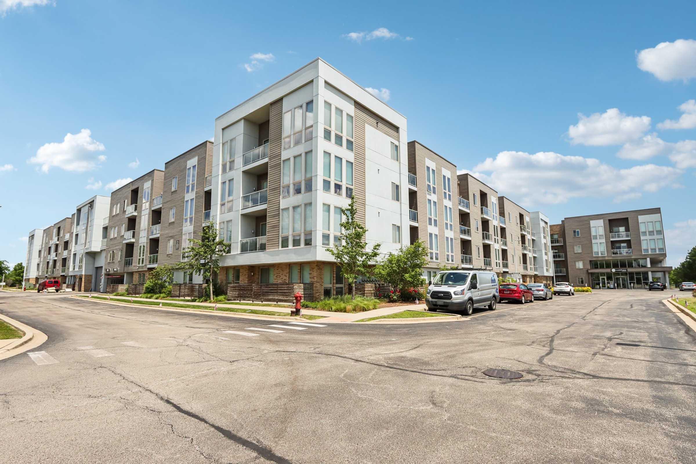 Modern apartment complex featuring three-story buildings with a mix of materials, large windows, and balconies. The scene includes a well-maintained street, a parked white van, a red car, and greenery, under a clear blue sky with scattered clouds.