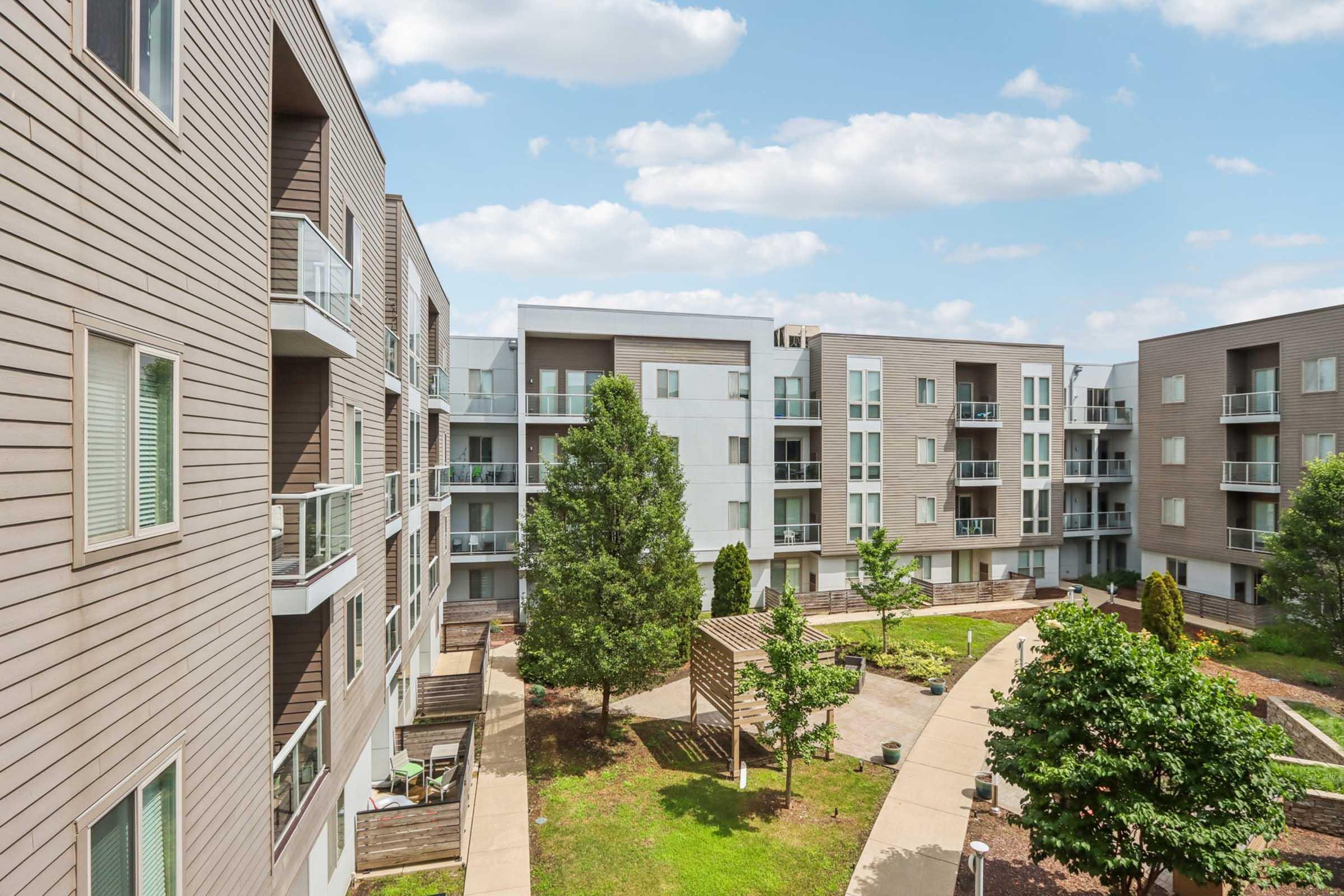An outdoor view of a modern apartment complex featuring several multi-story buildings with a mix of wood and light-colored facades. In the center, there is a landscaped courtyard with trees, green grass, and walking paths connecting the buildings under a partly cloudy sky.