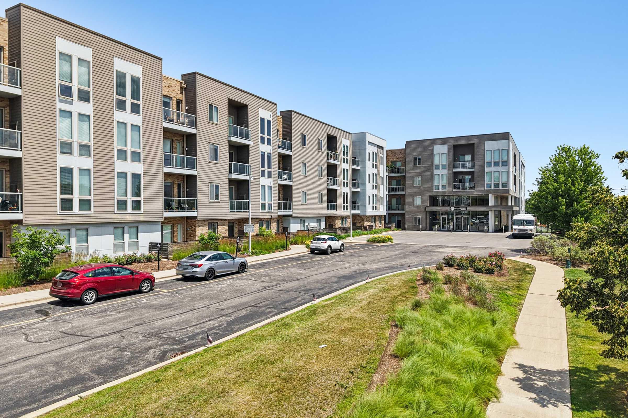 A modern apartment complex with multiple buildings featuring sleek designs and balconies. In the foreground, there is a paved road lined with parked cars and landscaped greenery. The setting is bright and open, with a clear blue sky above.