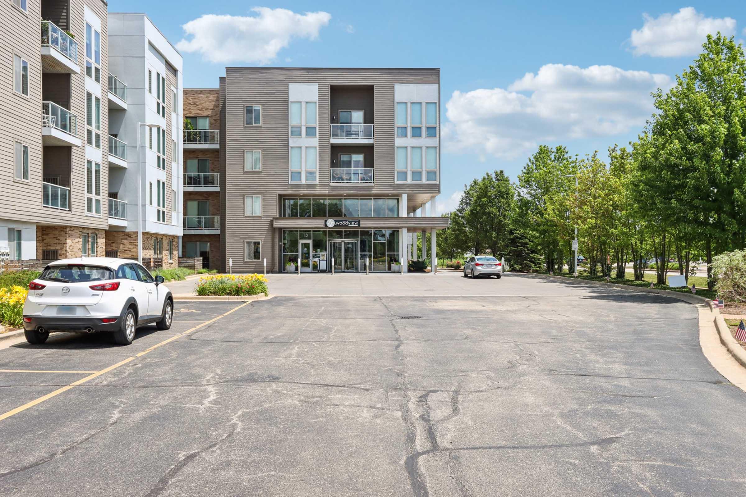 Modern apartment building with a glass entrance, surrounded by greenery and parked cars. The structure features a mix of wood and metal materials, and there is a clear blue sky with clouds in the background. The driveway is asphalt with clear parking lines.