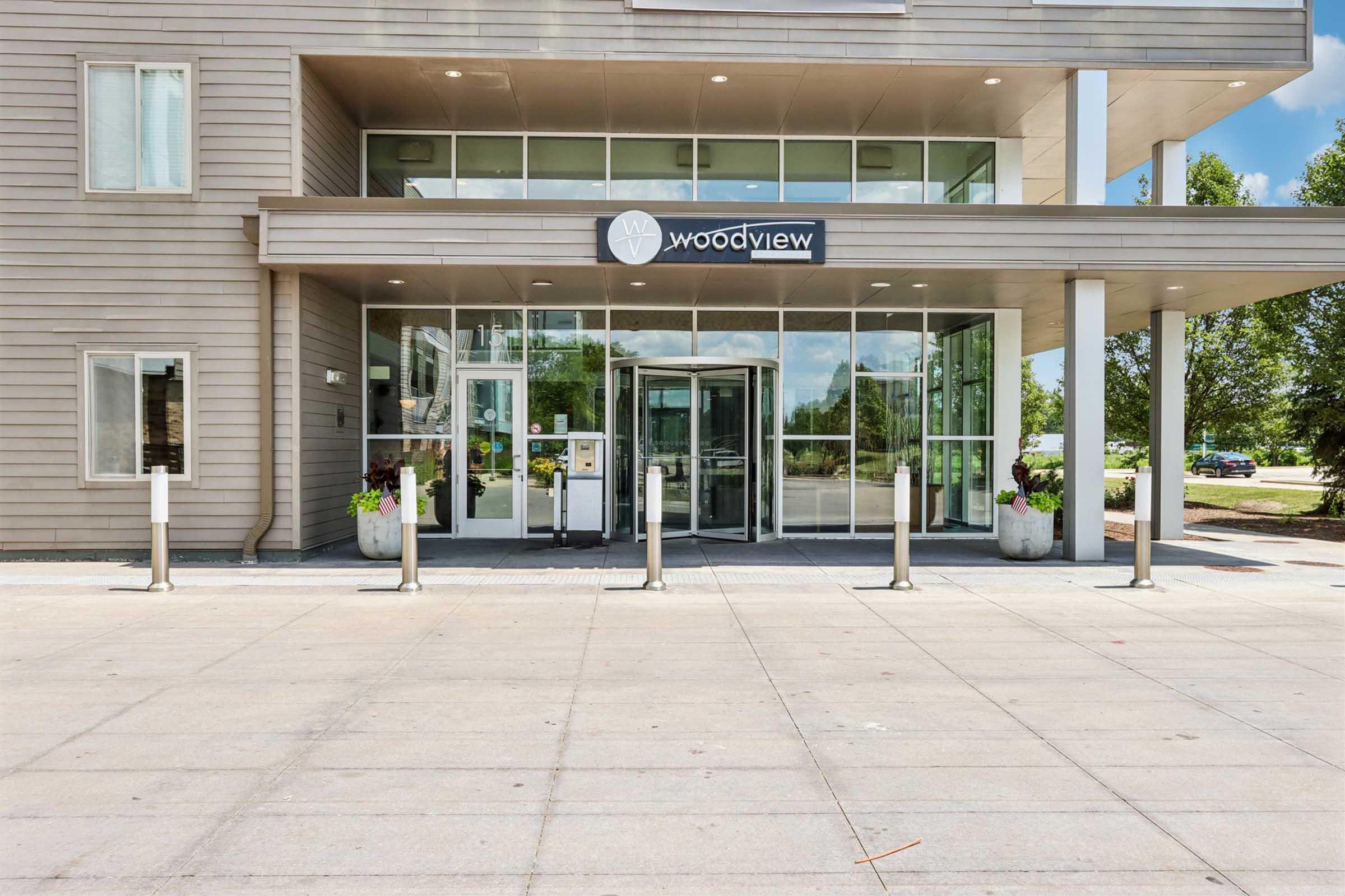 Entrance of a modern building named "woodview," featuring large glass doors and windows. The facade is finished in light wood and neutral tones, with potted plants on either side. There are metal bollards in front, and the area is well-lit with a clear blue sky in the background.