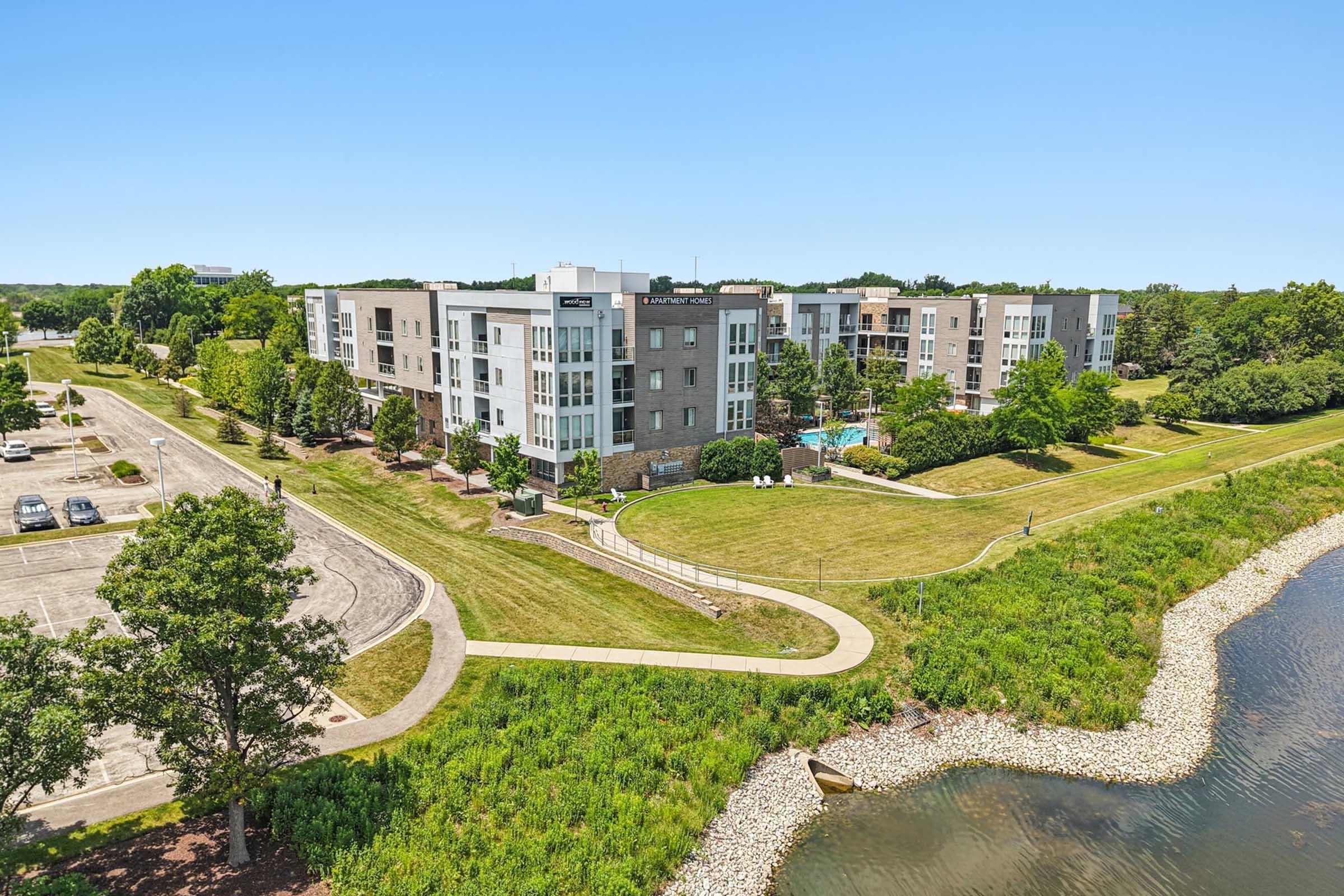 Aerial view of a modern apartment complex surrounded by green grass and trees, along a waterway. The building features balconies and large windows. In the foreground, there is a walking path and a parking lot nearby, indicating a well-maintained residential area. Clear blue sky overhead.