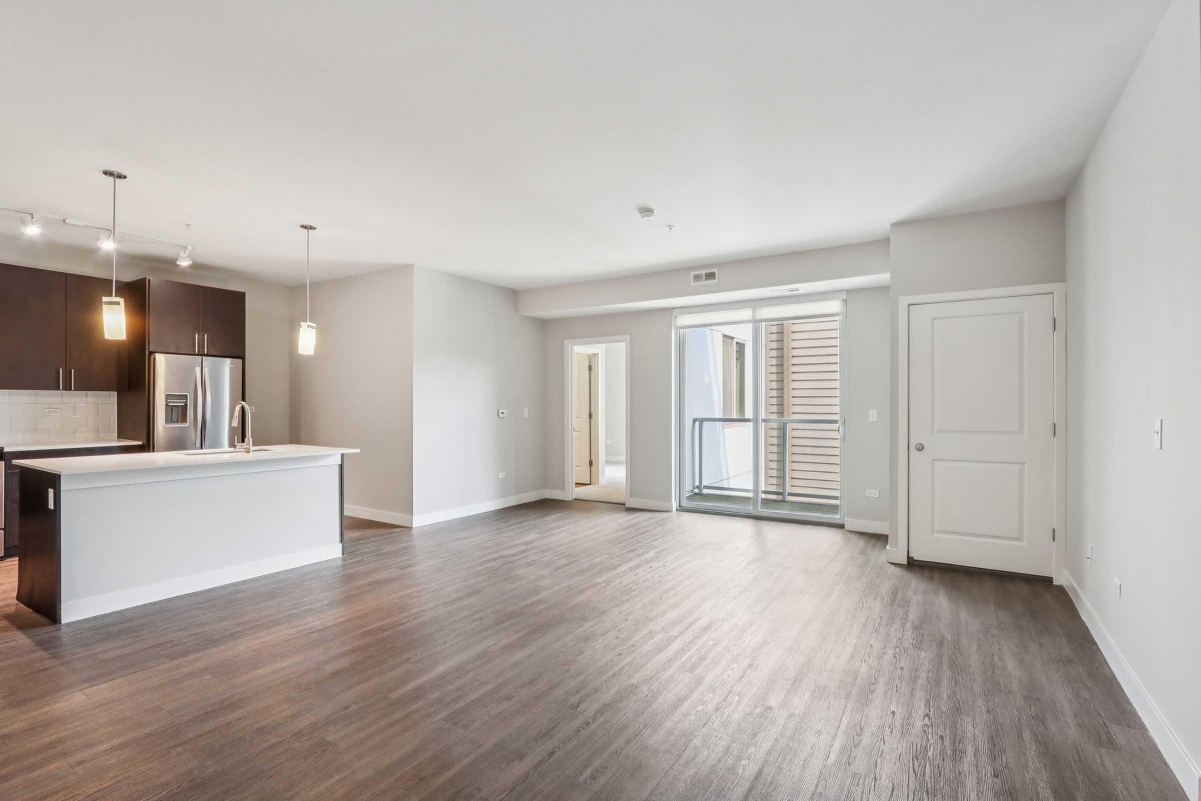 Spacious, modern apartment interior featuring an open floor plan with hardwood flooring. A kitchen area with a large island and stainless steel appliances is visible. Large windows provide natural light, leading to a balcony or outdoor space. Neutral wall colors create a clean and inviting atmosphere.