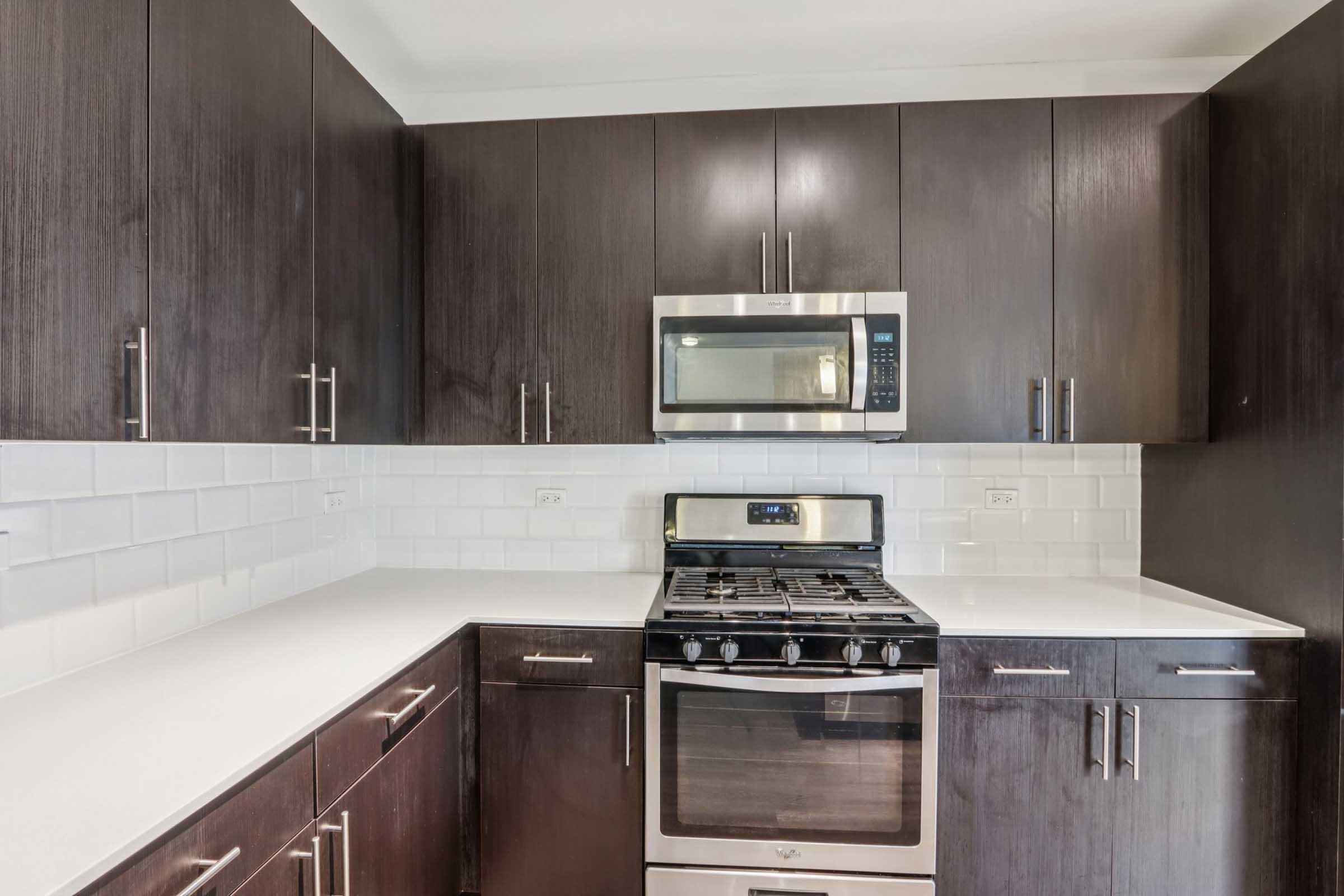 Modern kitchen with dark wood cabinets and a white countertop. It features a stainless steel microwave and gas range oven, complemented by white subway tile backsplash. The layout emphasizes a clean and contemporary design with sleek cabinet handles.