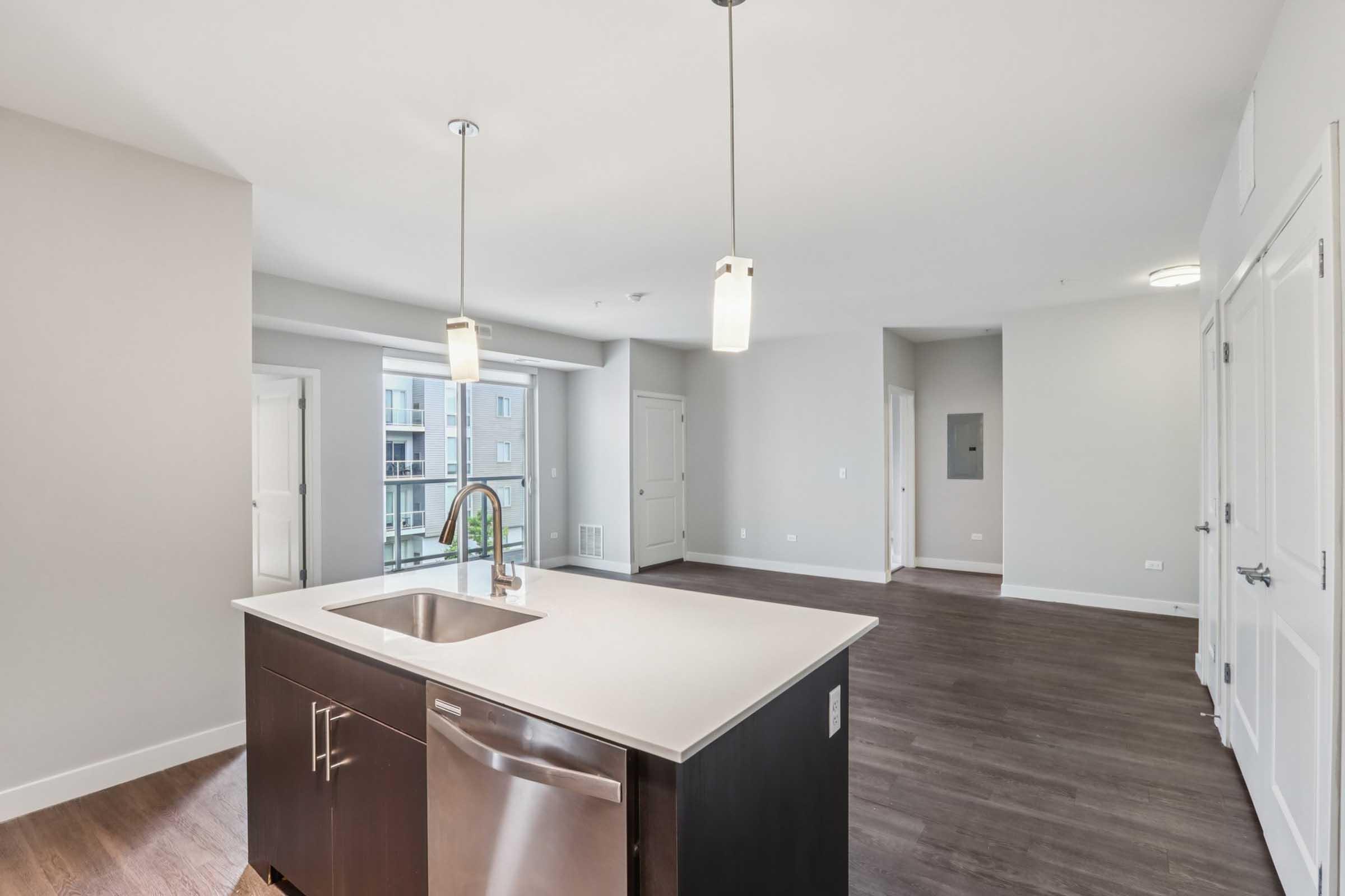 A modern kitchen with a central island featuring a stainless steel sink. Pendant lights hang above the island. The space has an open layout leading to a bright living area with large windows, light-colored walls, and hardwood flooring. Two doors lead to other rooms, enhancing the spacious feel.