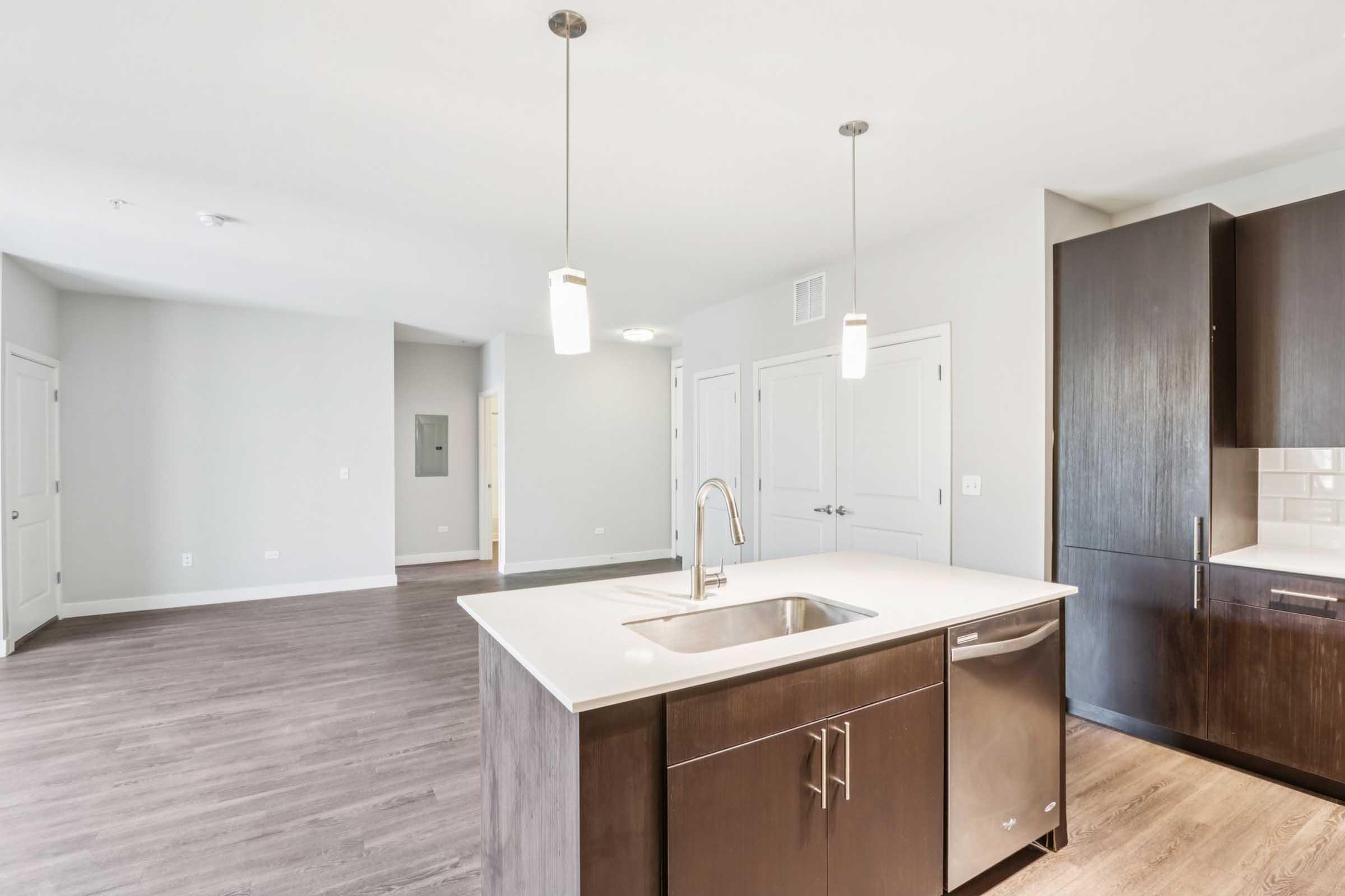 Modern kitchen with a central island featuring a sink, stainless steel dishwasher, and dark wood cabinetry. The open layout leads to a spacious living area with light gray walls and pendant lighting. Doors in the background indicate access to additional rooms or hallways.